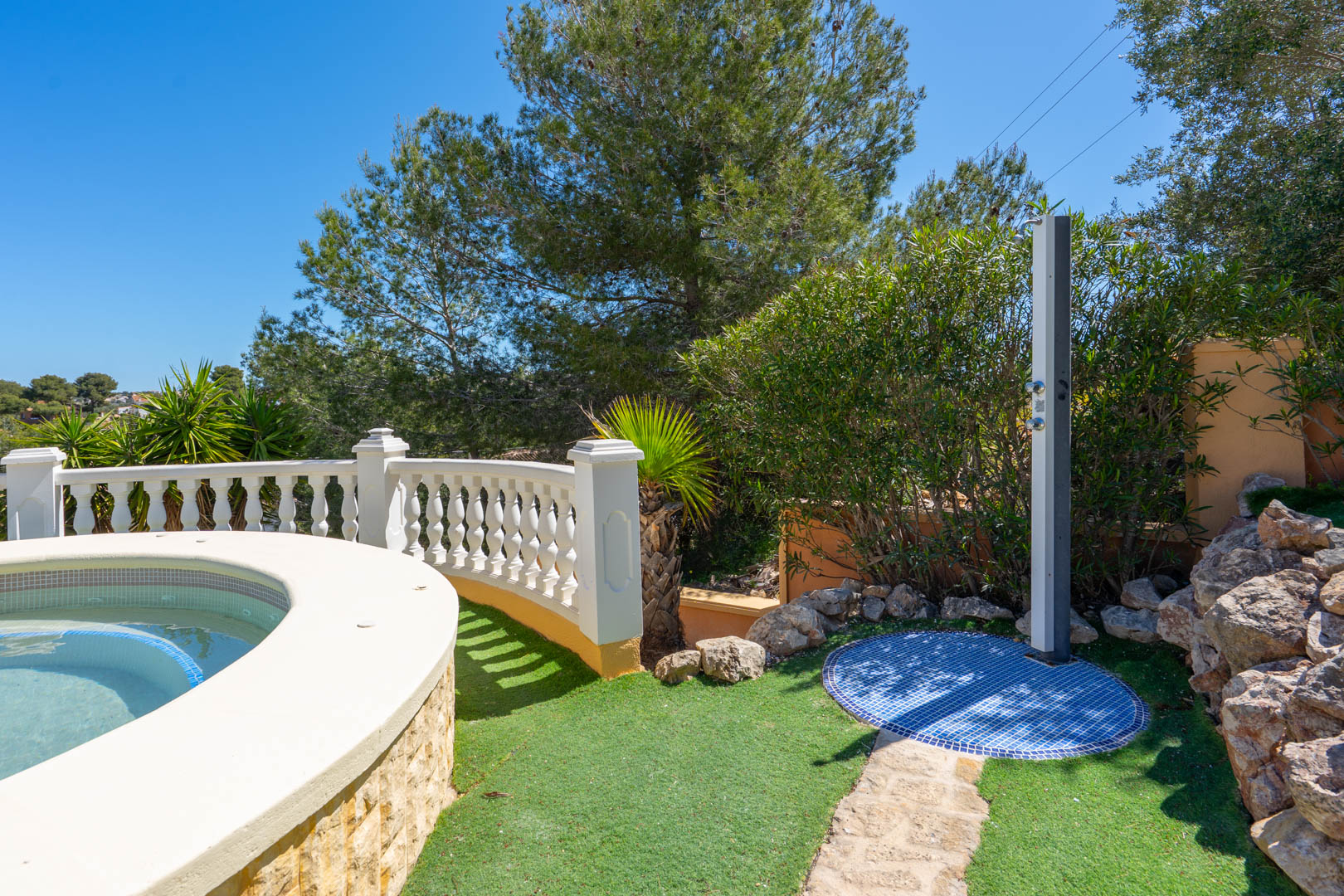 Außendusche mit blauem Mosaiksockel neben dem geschwungenen Poolrand, umgeben von einer weißen Balustrade und mediterraner Vegetation.