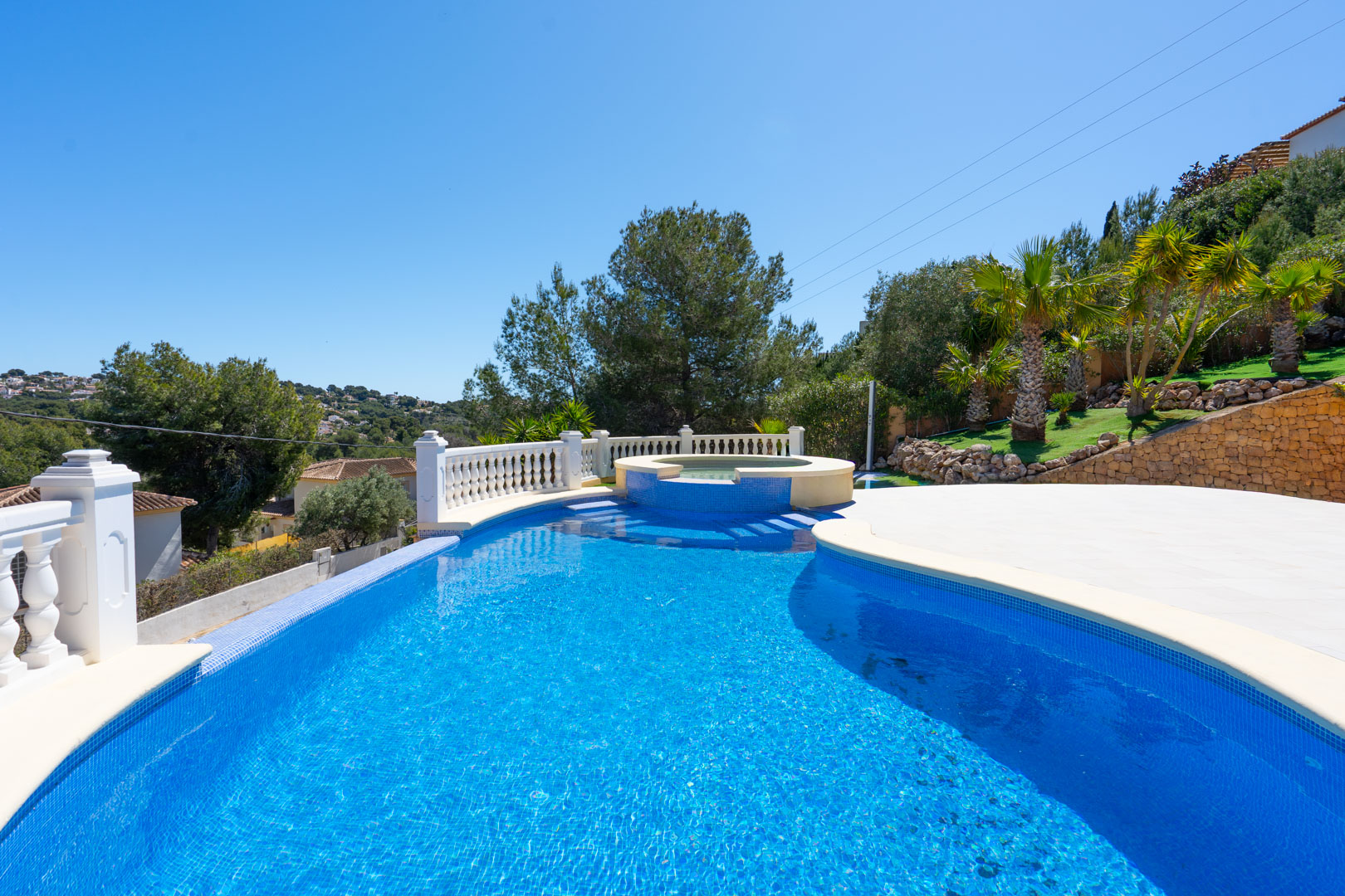Außenpool mit blauen Mosaikfliesen und integriertem Whirlpool, umgeben von einer Steinterrasse, weißer Balustrade und mediterraner Bepflanzung.