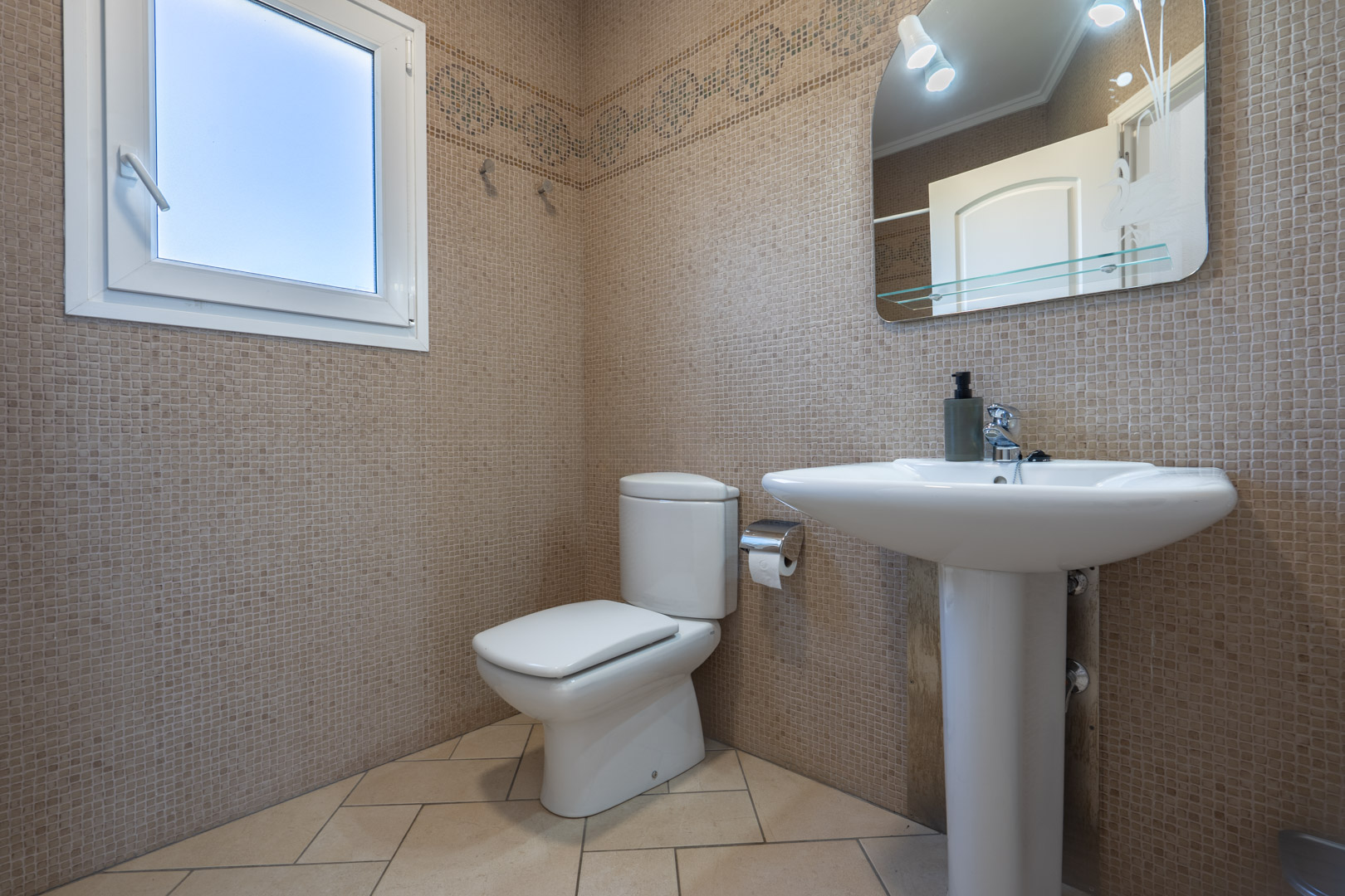 Badezimmer mit beigen Mosaikfliesen, einem Standwaschbecken und einem weißen PVC-Fenster für natürliches Tageslicht.