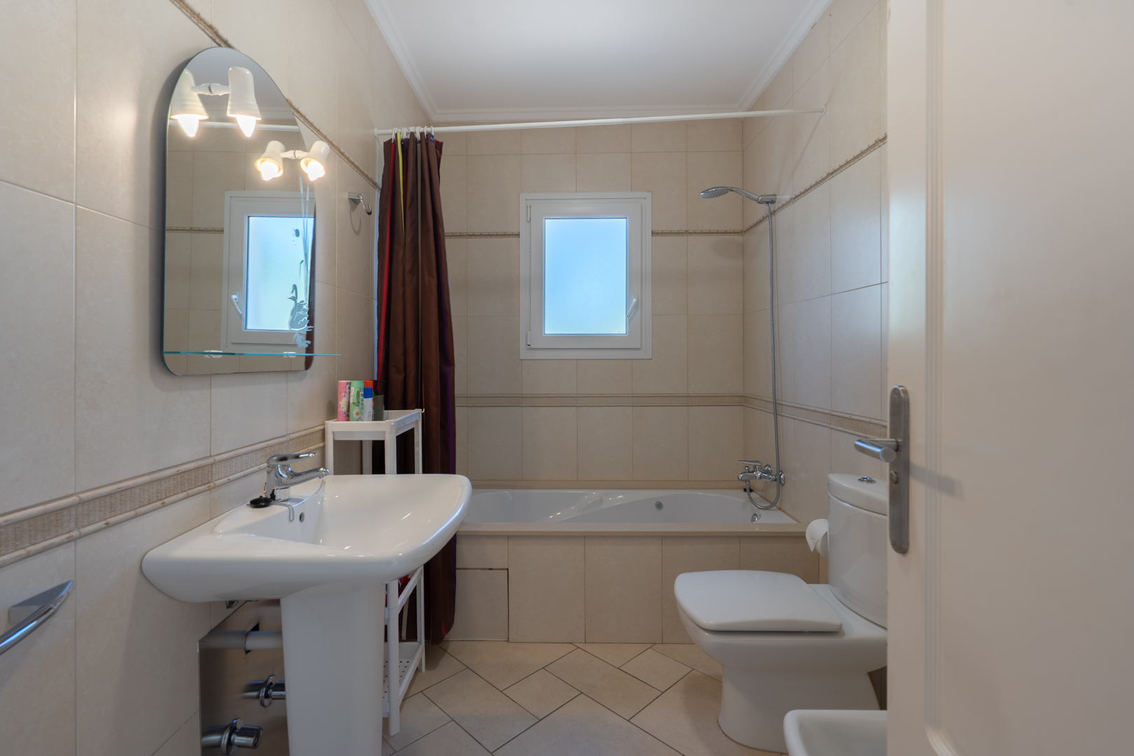 Badezimmer mit beigen Wandfliesen, Einbaubadewanne, Säulenwaschbecken und einem Bogenspiegel mit integrierter Beleuchtung neben einem PVC-Fenster.