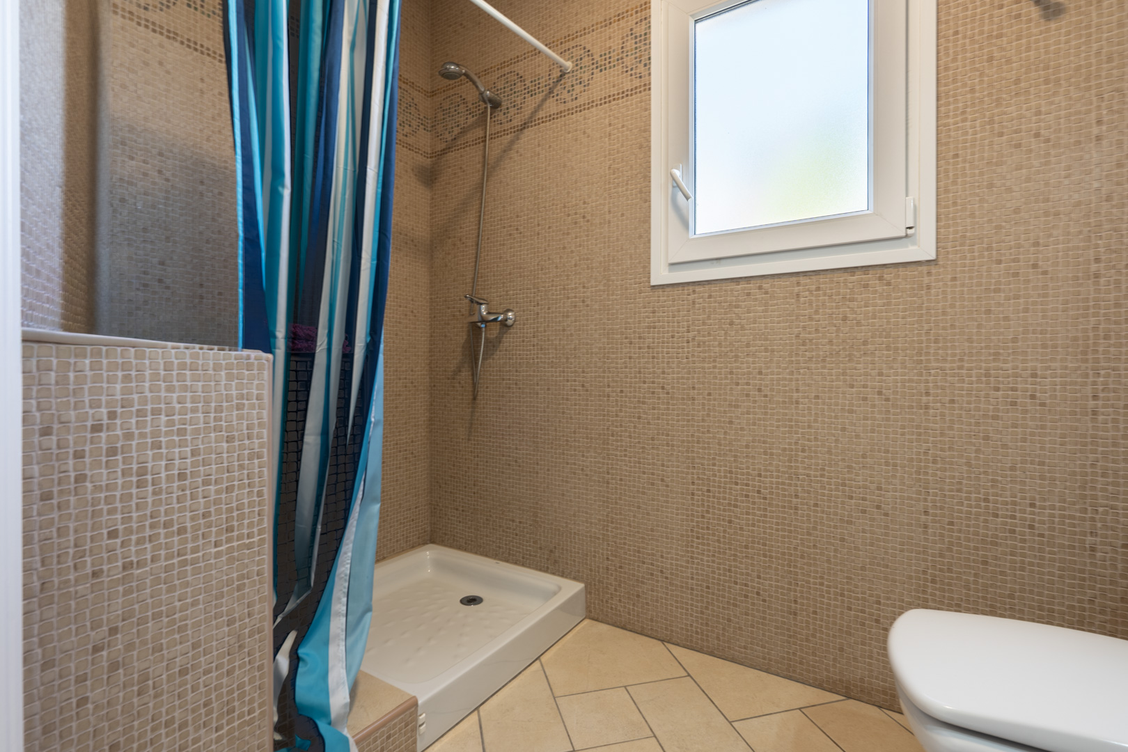 Badezimmer mit braunen Mosaikwänden, weißer Duschwanne mit gestreiftem Vorhang und einem weißen PVC-Fenster für Helligkeit und Belüftung.