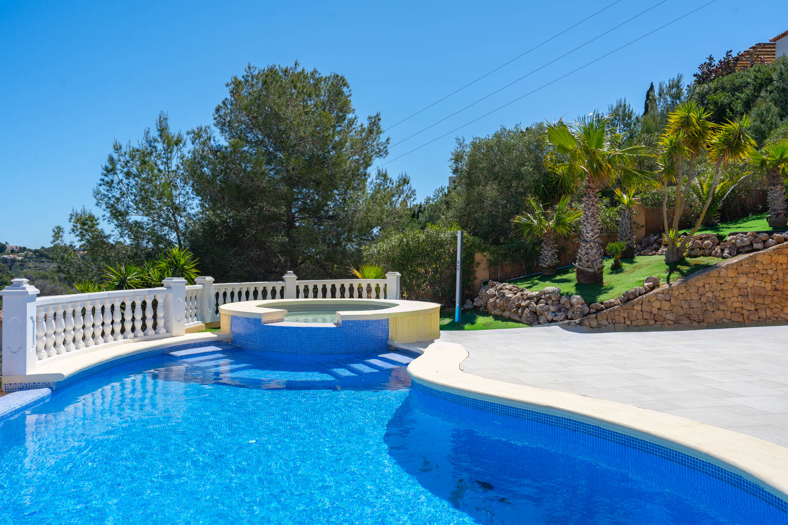 Ein 10x5 m großer Pool mit integriertem Whirlpool, blauen Mosaikfliesen, weißer Steinbalustrade und einem mediterranen Garten mit Palmen.