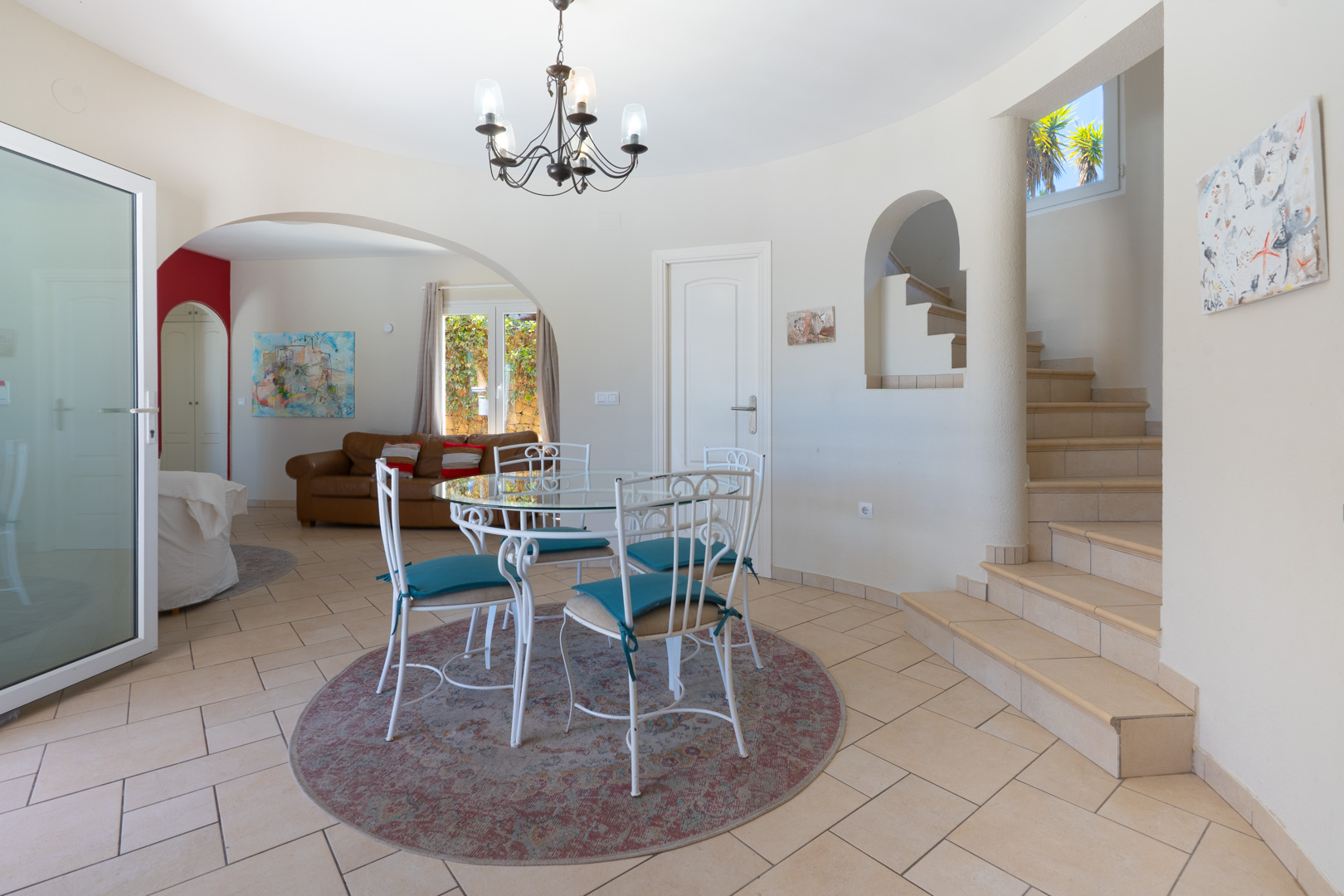 Essbereich mit Glastisch, weißen Schmiedeeisenstühlen und Kronleuchter. Der Raum bietet Zugang zu einer gefliesten Treppe und dem angrenzenden Wohnzimmer durch einen Bogen.