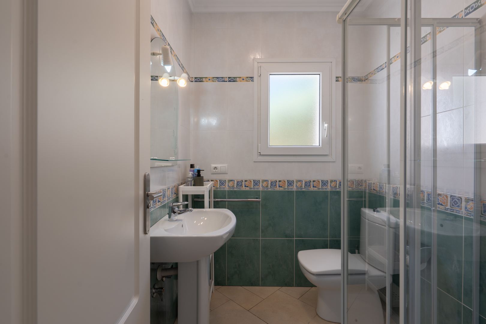 Funktionales Badezimmer mit grün-weißen Fliesen, Säulenwaschbecken, Glasduschkabine und einem PVC-Fenster mit Sichtschutzglas.
