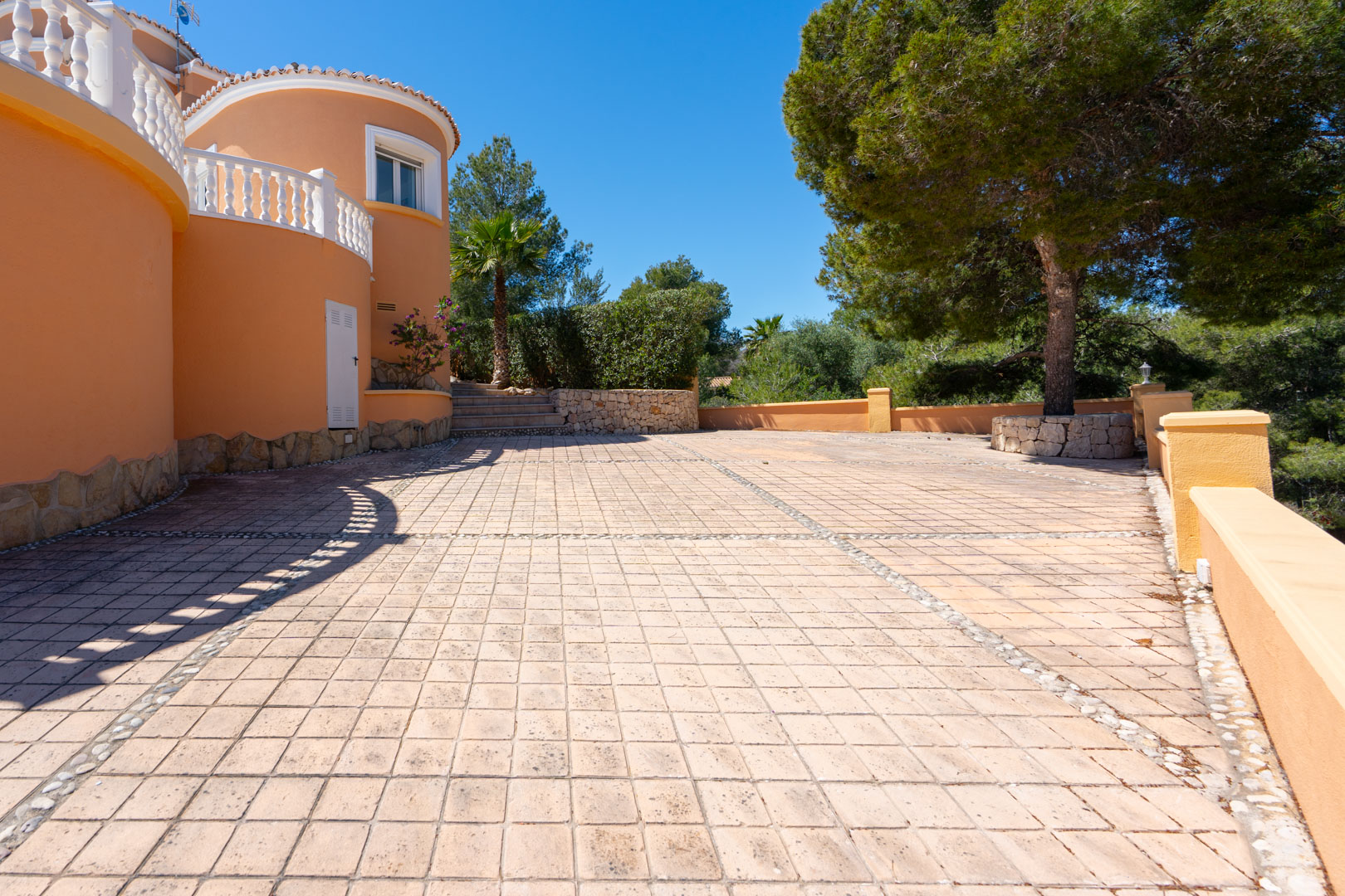 Großzügige gepflasterte Terrasse mit dekorativen Kieselsteinbordüren, geschwungenen terracottafarbenen Wänden und mediterranem Baumbestand.