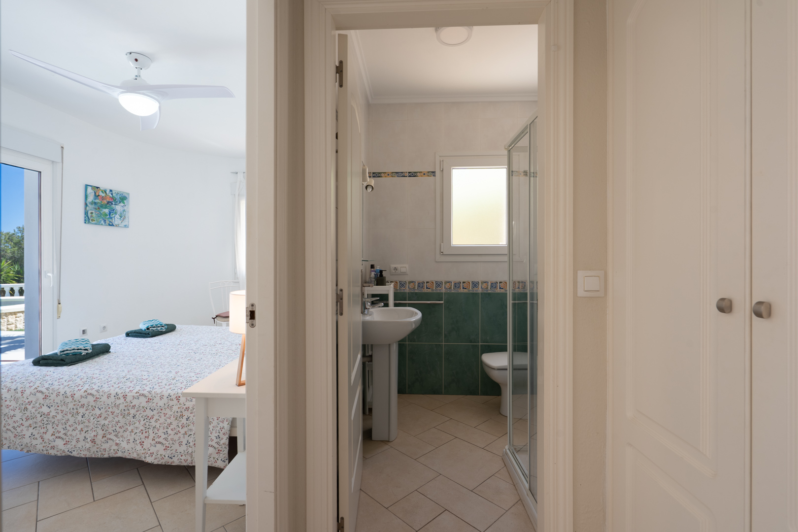 Helles Schlafzimmer mit Deckenventilator und direktem Außenzugang, neben einem Badezimmer mit Dusche und praktischen Einbauschränken im Flur.