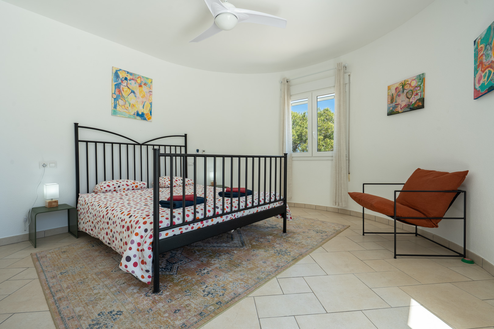 Helles Schlafzimmer mit geschwungener Wand, schwarzem Metallbettrahmen, Fliesenboden und PVC-Fenster mit Blick ins Grüne.