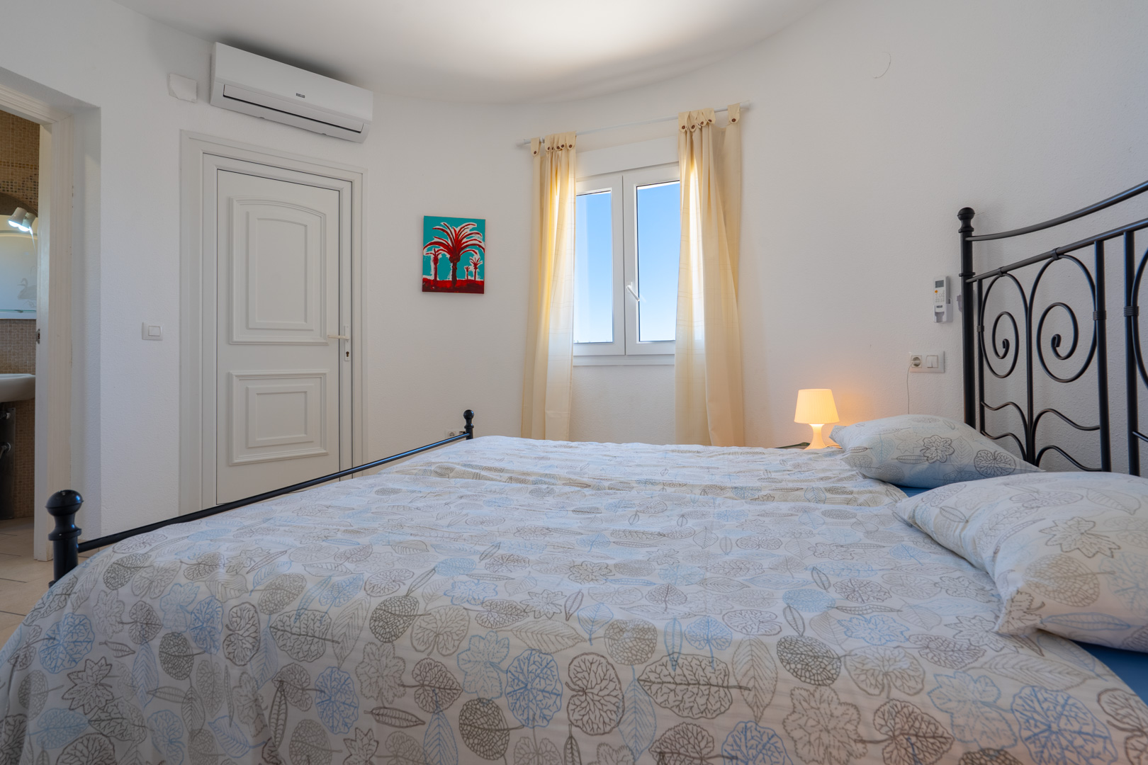 Helles Schlafzimmer mit schwarzem Schmiedeeisenbett, Klimaanlage, PVC-Fenster und direktem Zugang zum angrenzenden Badezimmer.