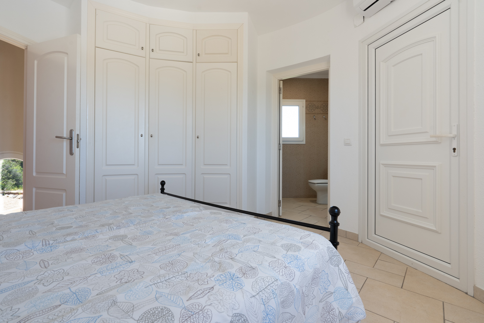Helles Schlafzimmer mit weißen Einbauschränken, Keramikfliesen und direktem Zugang zu einem angrenzenden Badezimmer mit Mosaikfliesen.