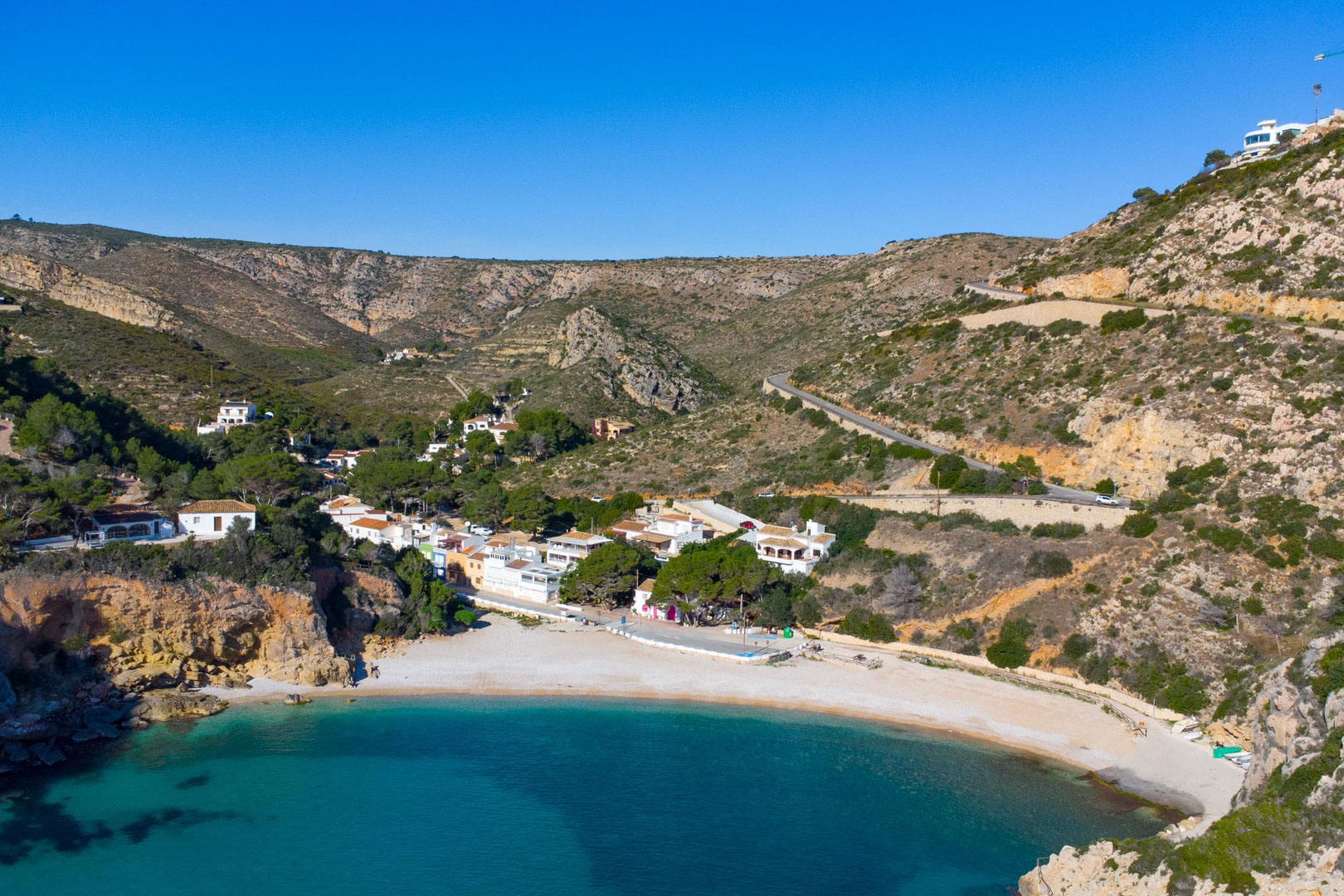Luftaufnahme der Cala Granadella in Jávea mit türkisblauem Wasser, Kieselstrand und den charakteristischen weißen Häusern in der felsigen Küstenlandschaft.