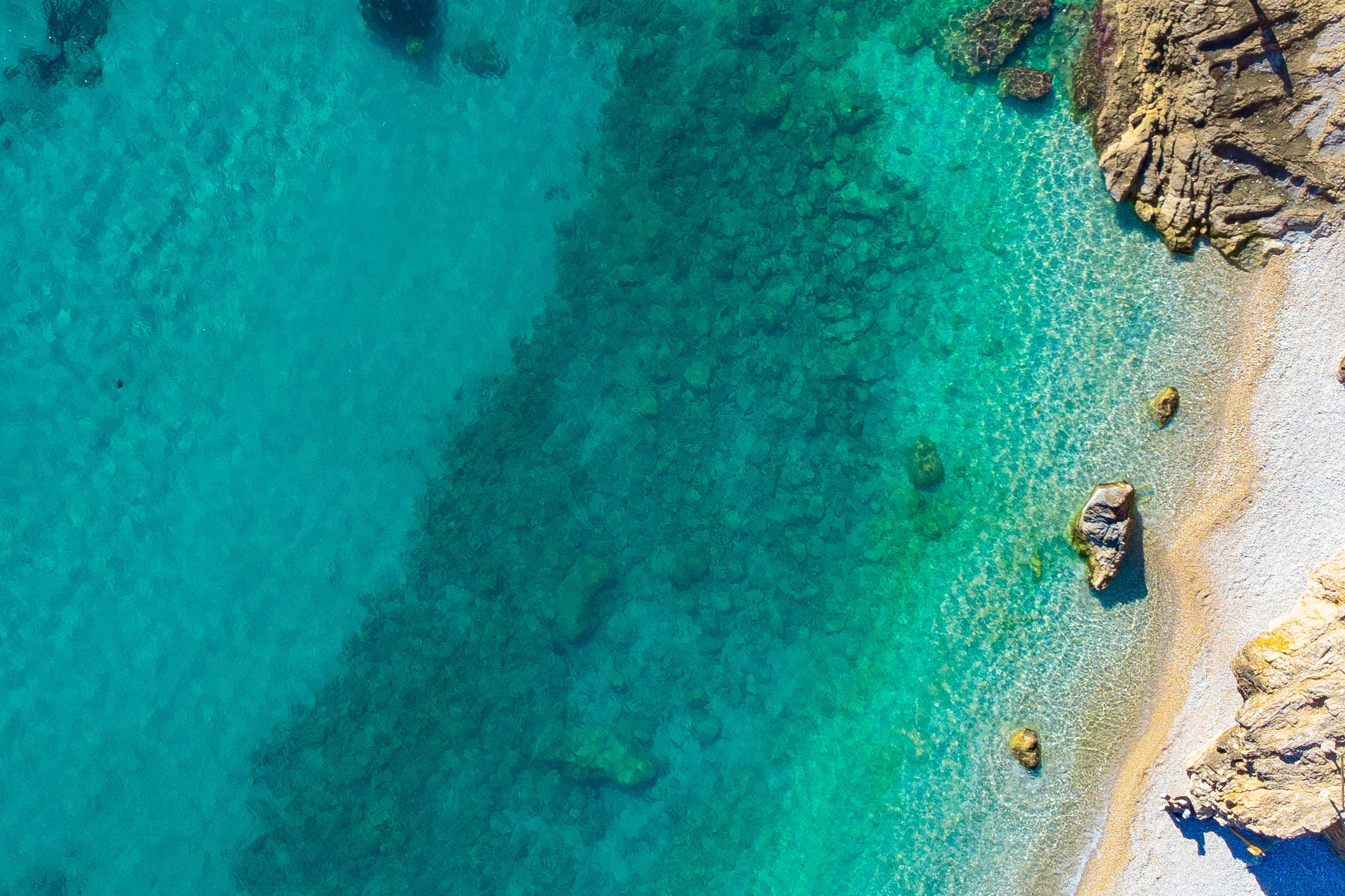 Luftaufnahme der Küste von Jávea mit kristallklarem, türkisfarbenem Wasser, sichtbarem felsigem Meeresboden und einem natürlichen Kieselstrand.