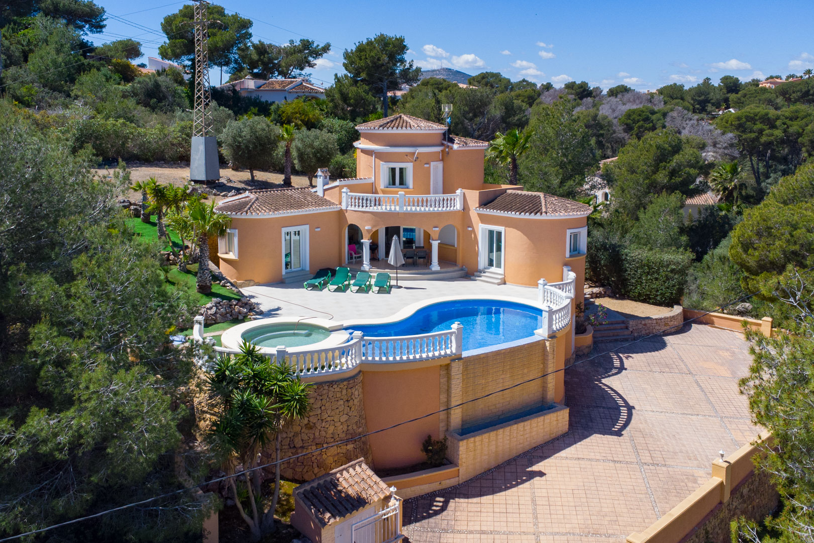 Luftaufnahme der mediterranen Villa mit geschwungenem Pool, integriertem Whirlpool und weißen Balustraden, umgeben von Pinien und einer gepflasterten Einfahrt.