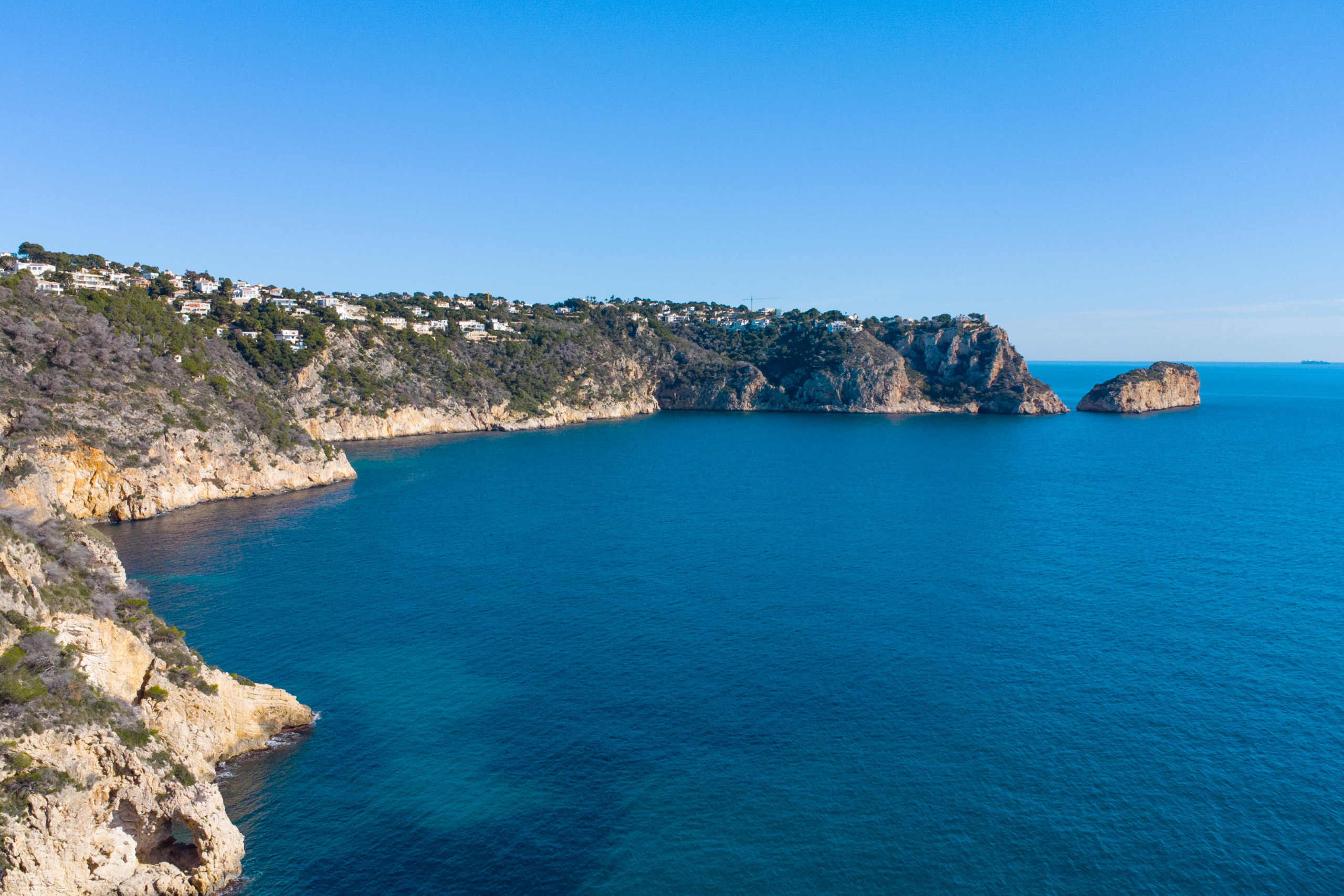 Panoramablick auf die Steilküste von Jávea mit markanten Kalksteinfelsen, dem tiefblauen Mittelmeer und Wohnvillen in exponierter Küstenlage.