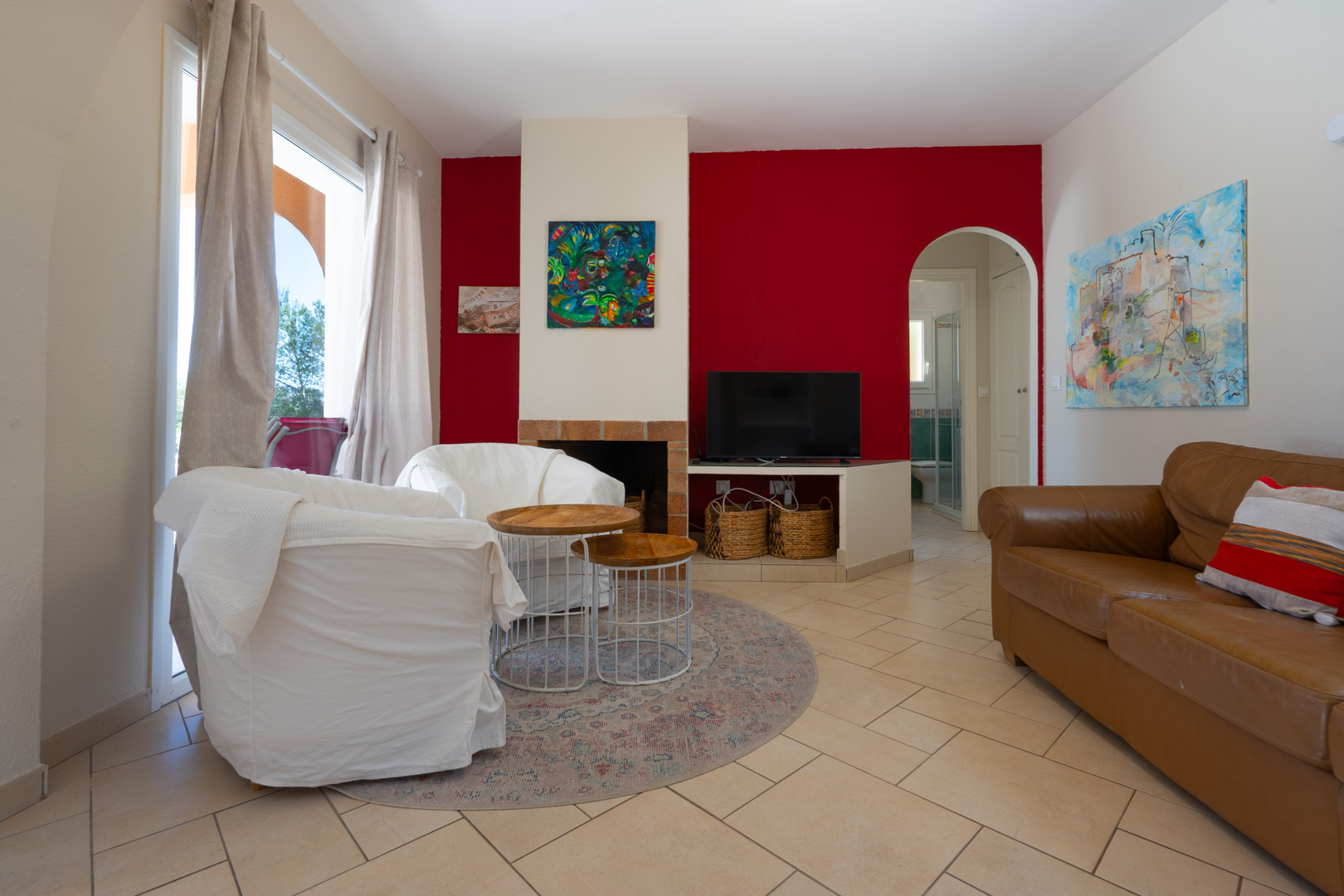 Wohnbereich mit roter Akzentwand, eingebautem Kamin und Rundbogen. Ausgestattet mit Fliesenboden, Ledersofa und großen Fenstern für viel Tageslicht.