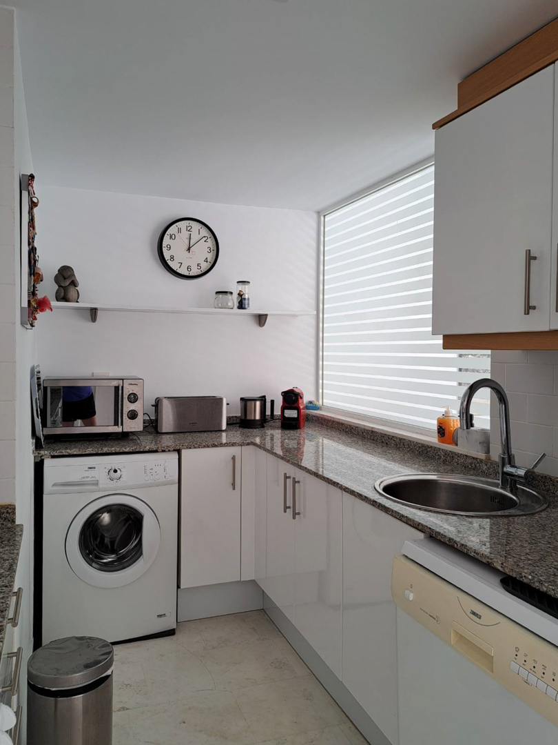 Funktionale L-Küche mit Granitarbeitsplatte, weißen Schränken, integrierter Waschmaschine und einem großen Fenster für optimalen Lichteinfall.