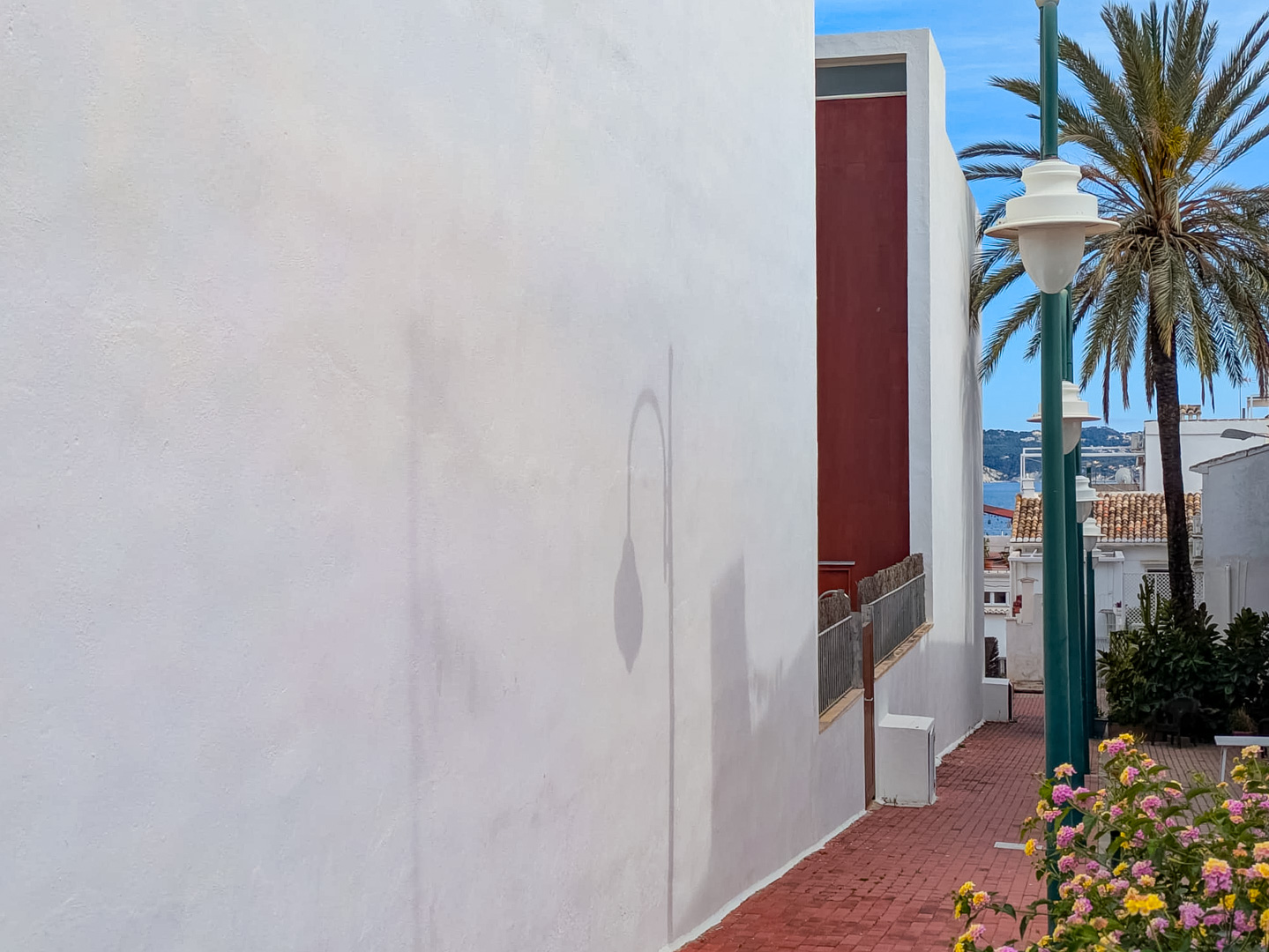 Gebäudeaußenansicht mit weißer Fassade und einem gepflasterten Fußweg, der Teilblicke auf das Meer und mediterrane Pflanzen bietet.