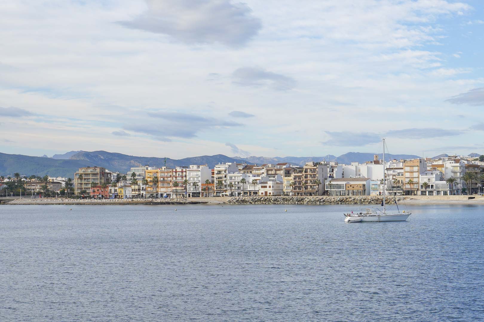 Panoramablick auf die Küste des Hafens von Jávea vom Meer aus, mit der Architektur der ersten Meereslinie, einem Segelboot und den Bergen im Hintergrund.