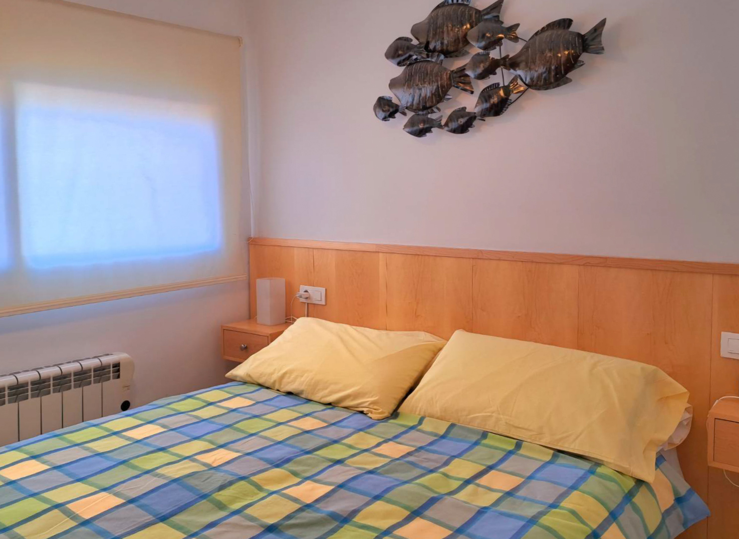 Schlafzimmer mit integriertem Holzkopfteil, passenden Nachttischen und Wanddekoration aus Metall. Ausgestattet mit Elektroheizkörper und Fenster mit Rollo.