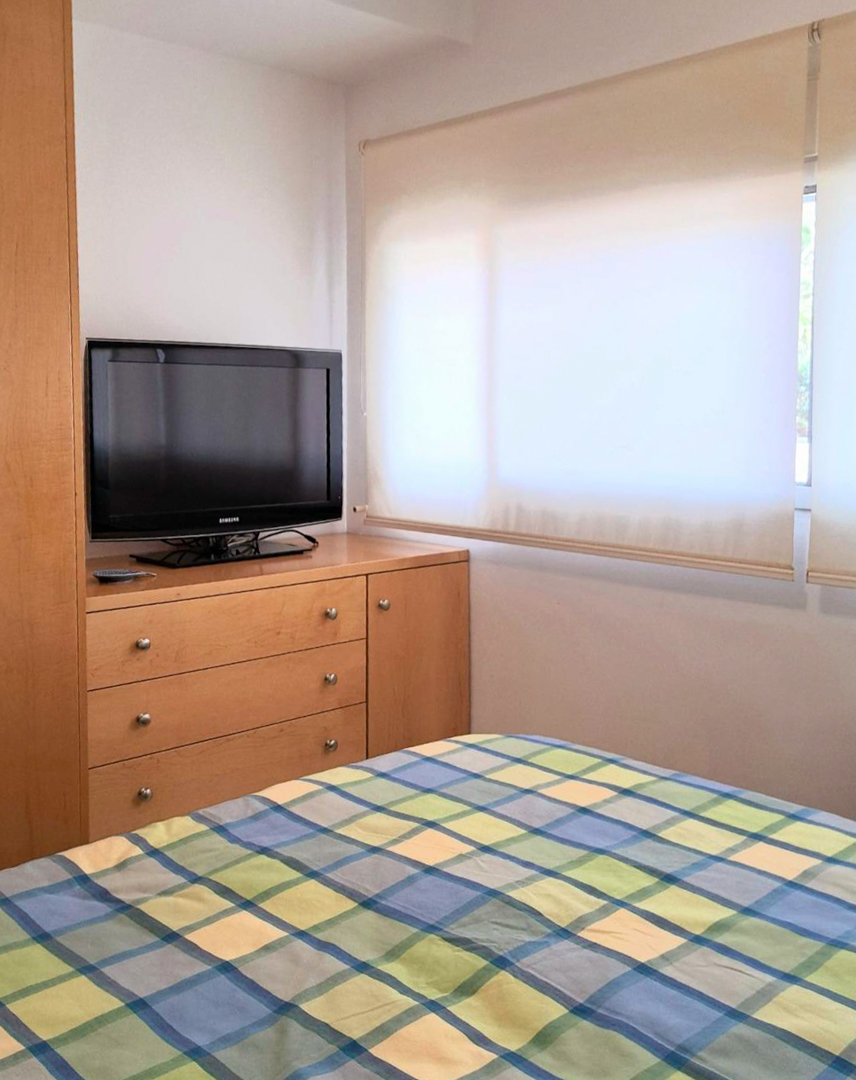 Schlafzimmer mit maßgefertigten Holzmöbeln, Fernseher auf einer Kommode und Fenster mit lichtdurchlässigen Rollos für natürliches Licht.