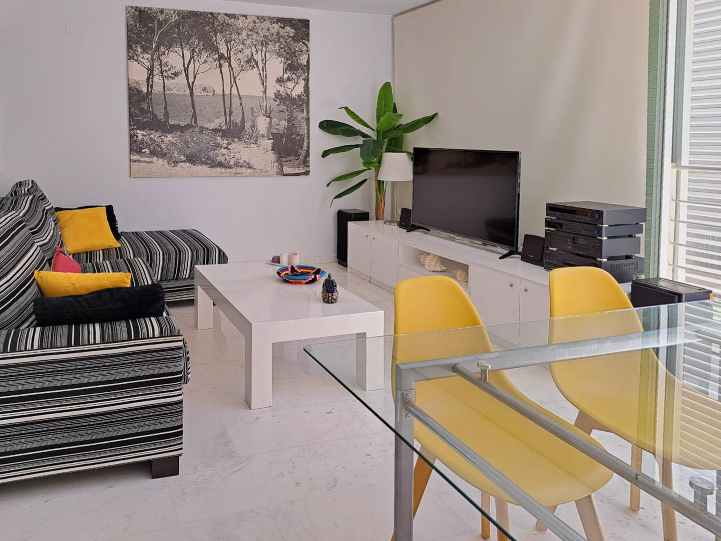 Wohn-Essbereich mit Marmorboden, gestreiftem Sofa und Glastisch mit gelben Stühlen. Ein weißes TV-Board und große Wandkunst ergänzen das funktionale Design.