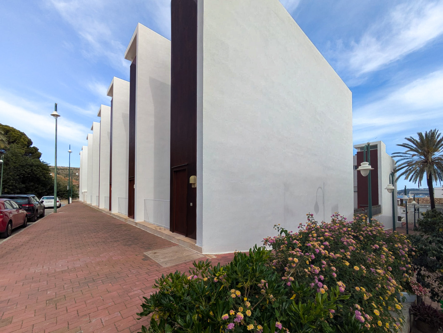 Zeitgenössische Außenfassade mit weißen Wänden und dunklen vertikalen Elementen, gelegen an einem gepflasterten Weg mit blühenden mediterranen Sträuchern.