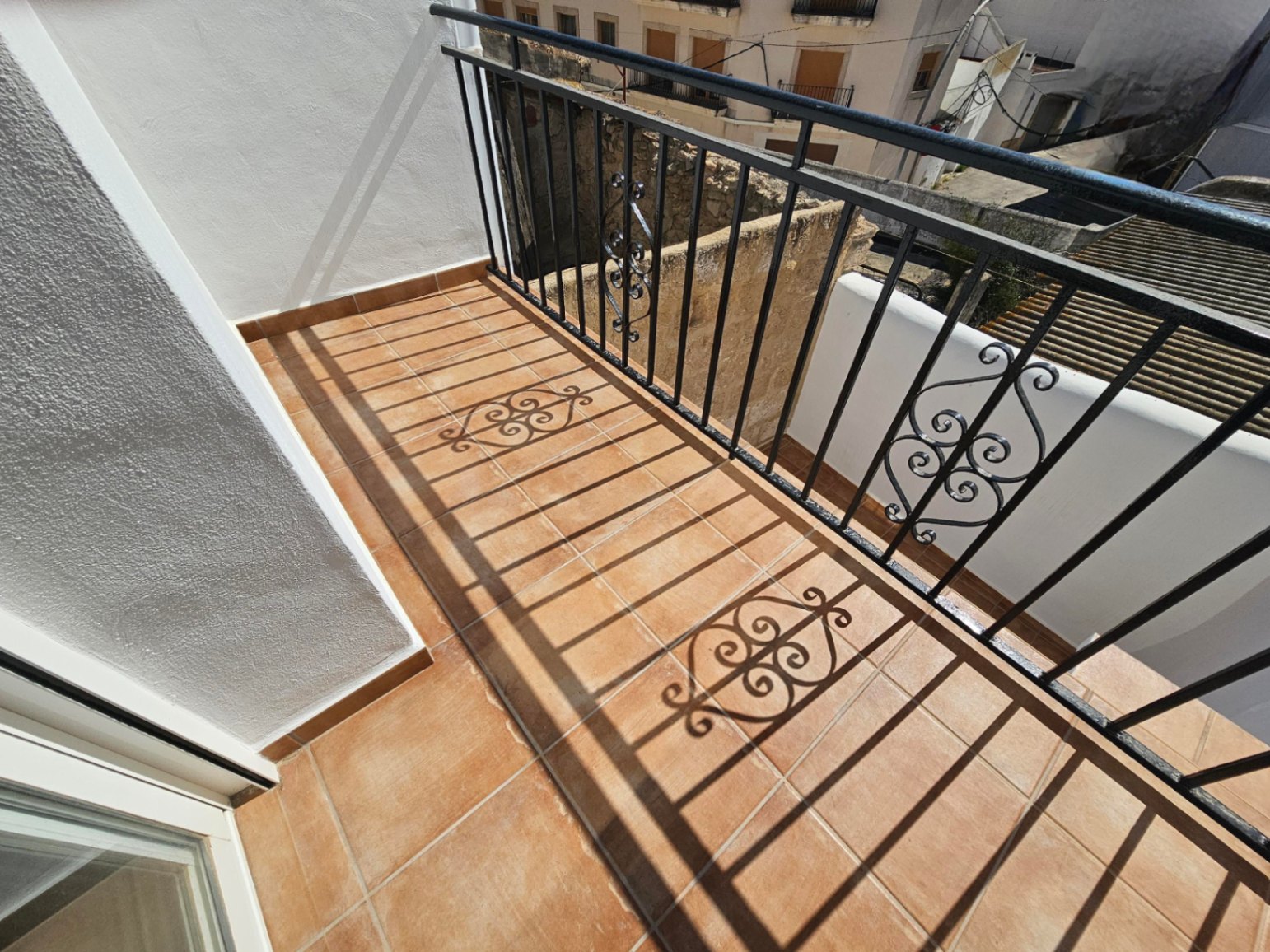 Balkon mit Terrakotta-Fliesenboden und schwarzem Schmiedeeisengeländer mit dekorativen Ornamenten, mit Blick auf die traditionelle Dorfumgebung.