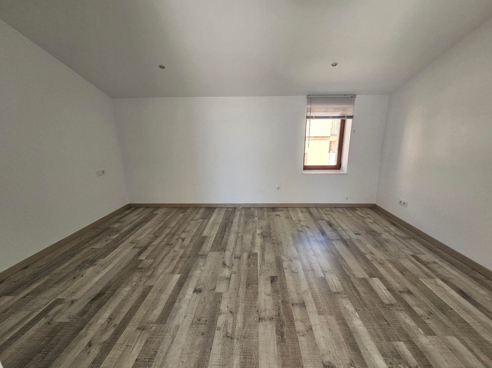 Heller Dachraum mit Dachschrägen, grauem Laminatboden und Deckeneinbaustrahlern. Ein Holzfenster mit Jalousie sorgt für natürliches Licht in diesem modernisierten Zimmer.