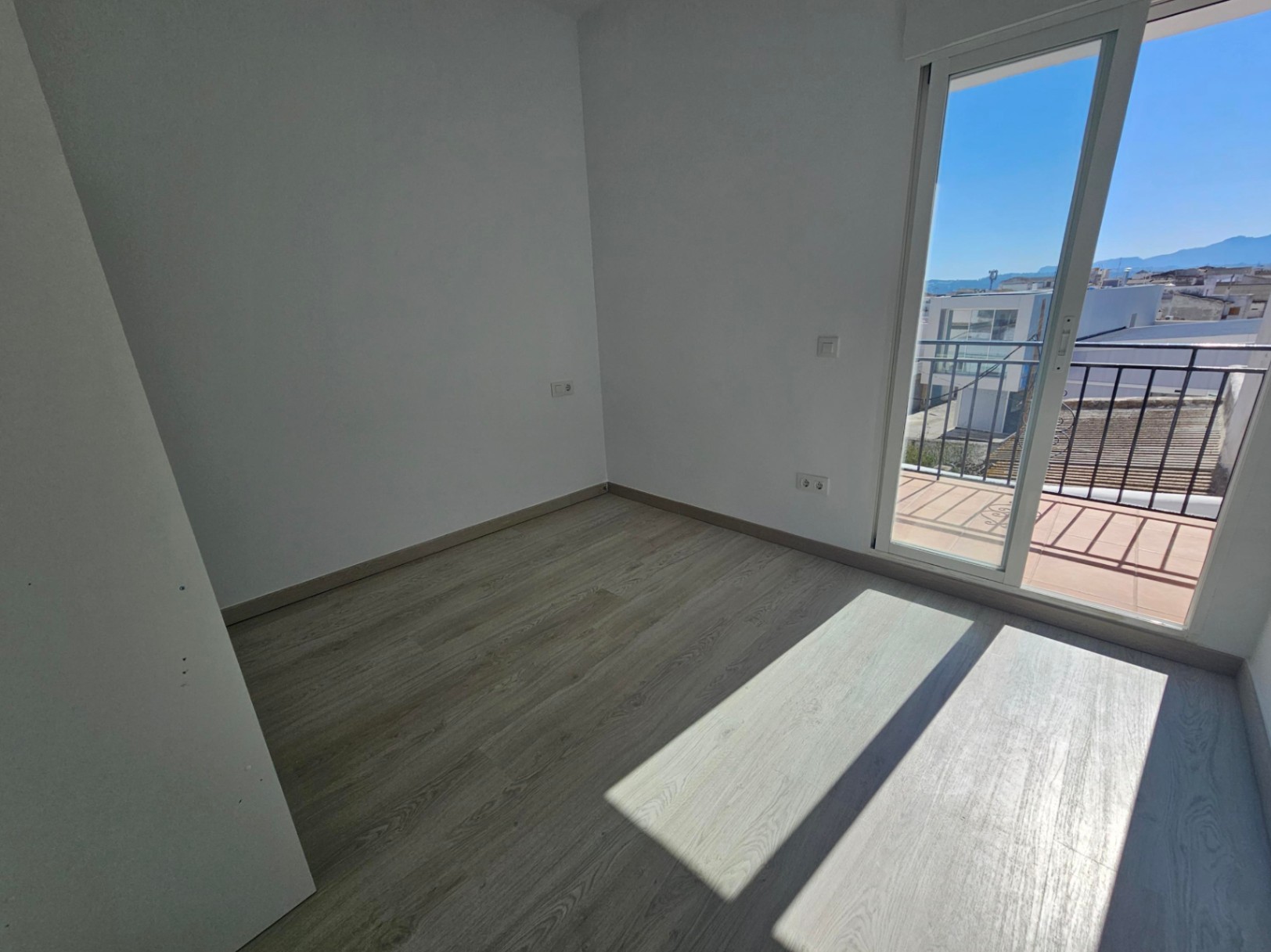 Helles Schlafzimmer mit hellem Laminatboden, weißen Wänden und Glasschiebetür zu einem privaten Balkon mit weitem Blick auf das Dorf und die umliegenden Berge.
