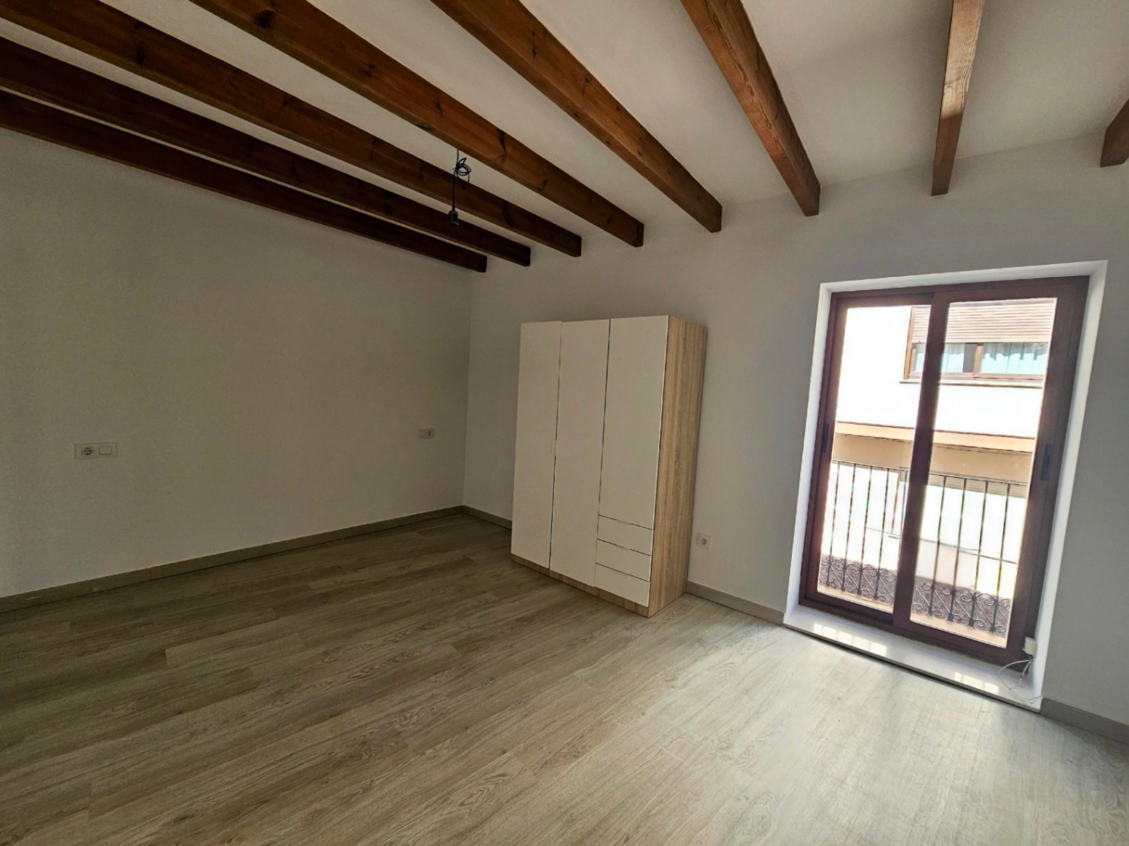 Schlafzimmer mit sichtbaren Holzbalken, hellem Laminatboden und direktem Zugang zu einem Balkon durch großzügige Schiebetüren.