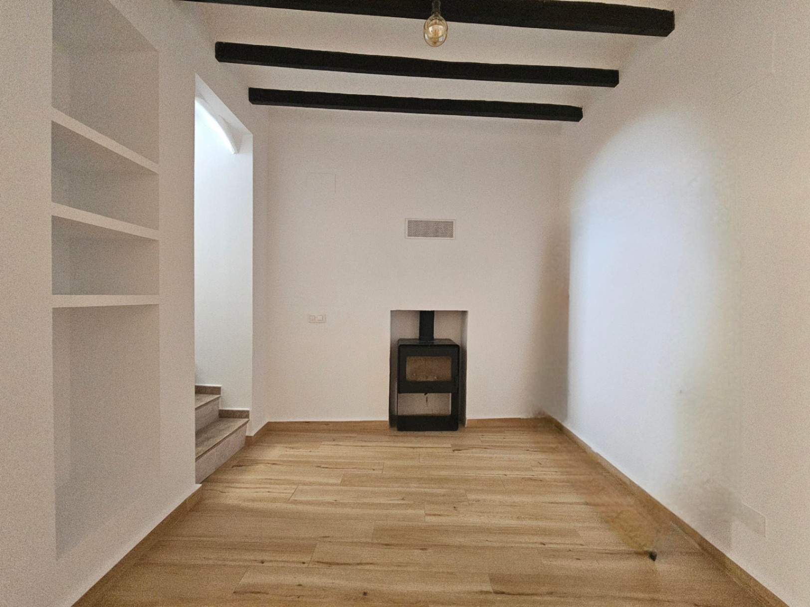 Wohnbereich mit dunklen Deckenbalken, eingebautem Regal und einem Kaminofen. Heller Boden in Holzoptik und weiße Wände sorgen für ein modernes Ambiente.