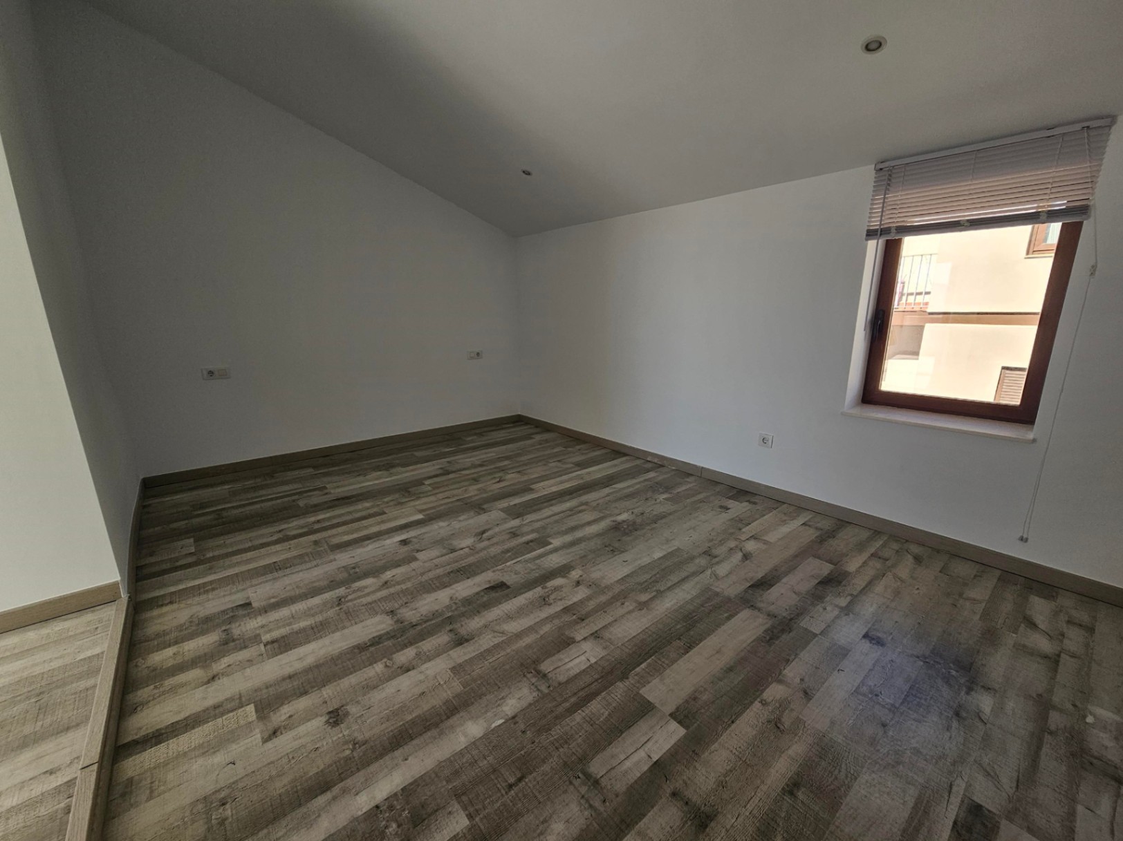 Zimmer mit Dachschräge, Laminatboden in Holzoptik und Einbaustrahlern. Ein Holzfenster mit Jalousie sorgt für helles, natürliches Licht.