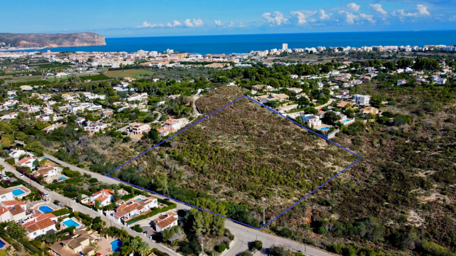 Luftaufnahme des 17.272 m² großen Grundstücks in Jávea, blau umrandet, in einem Wohngebiet mit Blick auf das Mittelmeer und die Küstenlinie gelegen.