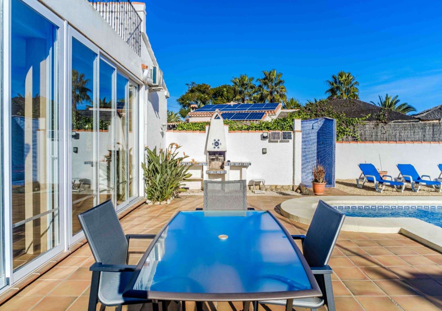 Außenterrasse mit Glastisch, gemauertem Grill und blau gefliester Dusche am Pool. Große Glasschiebetüren und Solarmodule sorgen für Funktionalität und Komfort.
