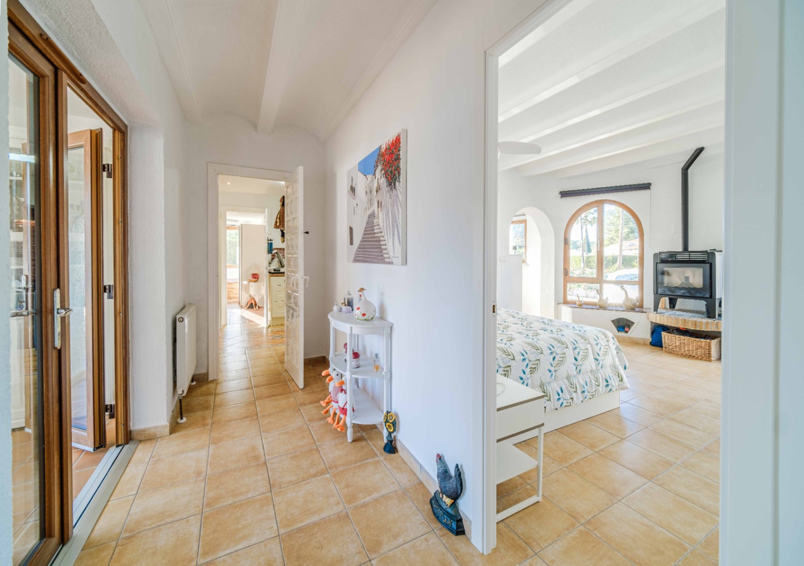Flur mit Terrakotta-Fliesen, der in ein helles Schlafzimmer mit weißen Deckenbalken, Rundbogenfenster und einem freistehenden Kaminofen führt.