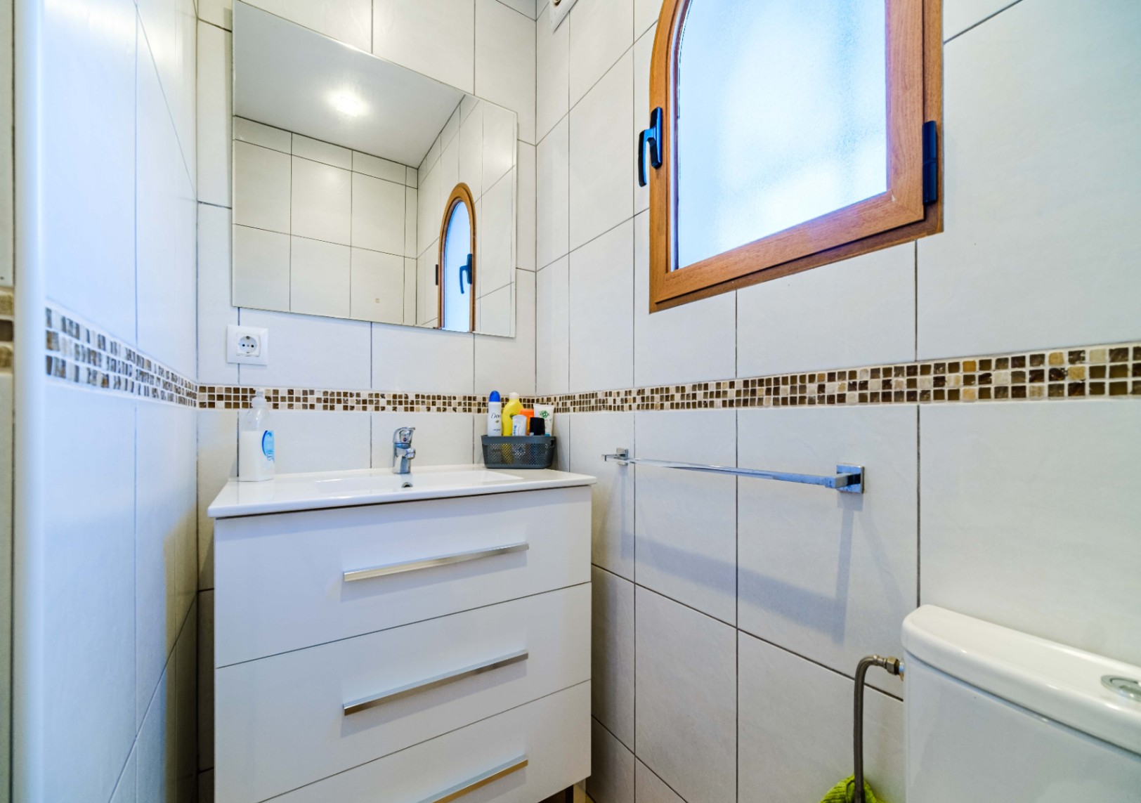 Funktionales Badezimmer mit weißem Waschtisch mit drei Schubladen, rechteckigem Spiegel und gewölbtem Holzfenster. Geflieste Wände mit Mosaikbordüre.