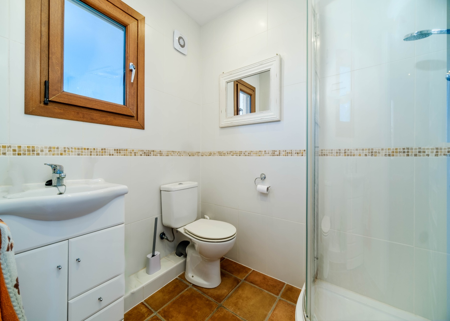 Funktionales Badezimmer mit weißen Fliesenwänden, Mosaikbordüre, Terrakottaboden und einer Glasduschkabine.