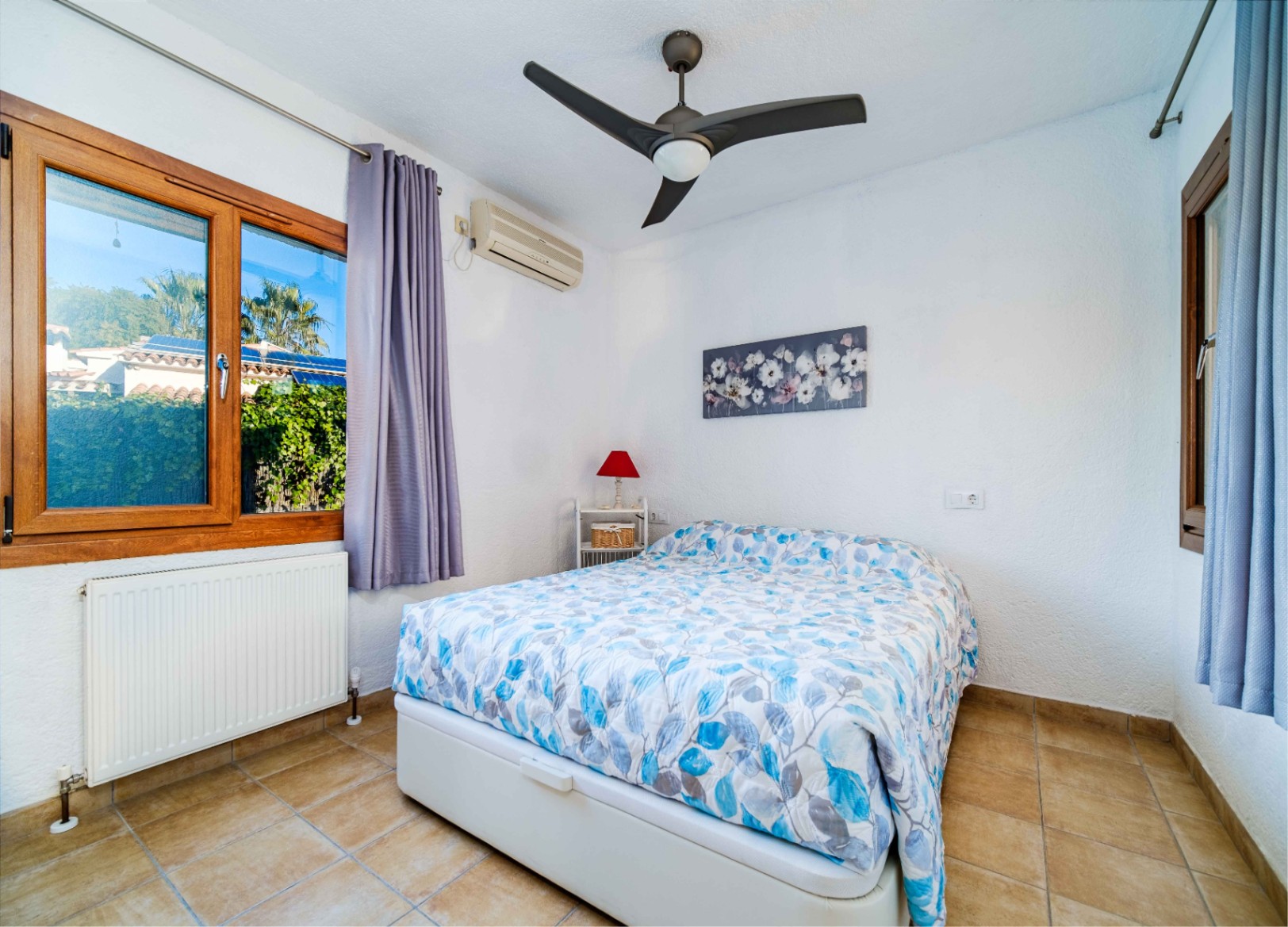 Helles Schlafzimmer mit Fliesenboden, Deckenventilator, Klimaanlage und Holzfenstern mit Blick auf die gepflegte Gartenanlage.