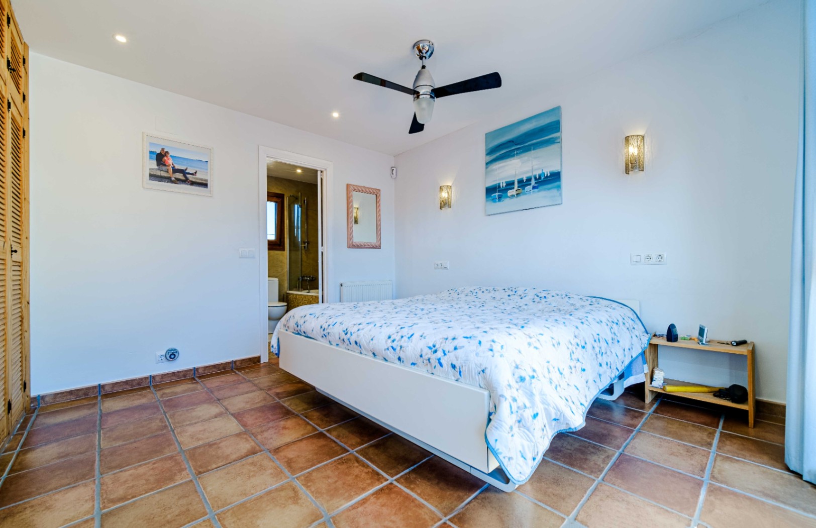 Helles Schlafzimmer mit rustikalen Fliesenböden, Deckenventilator und En-Suite-Badezimmer. Ausgestattet mit Einbauschränken aus Holz und Wandleuchten.