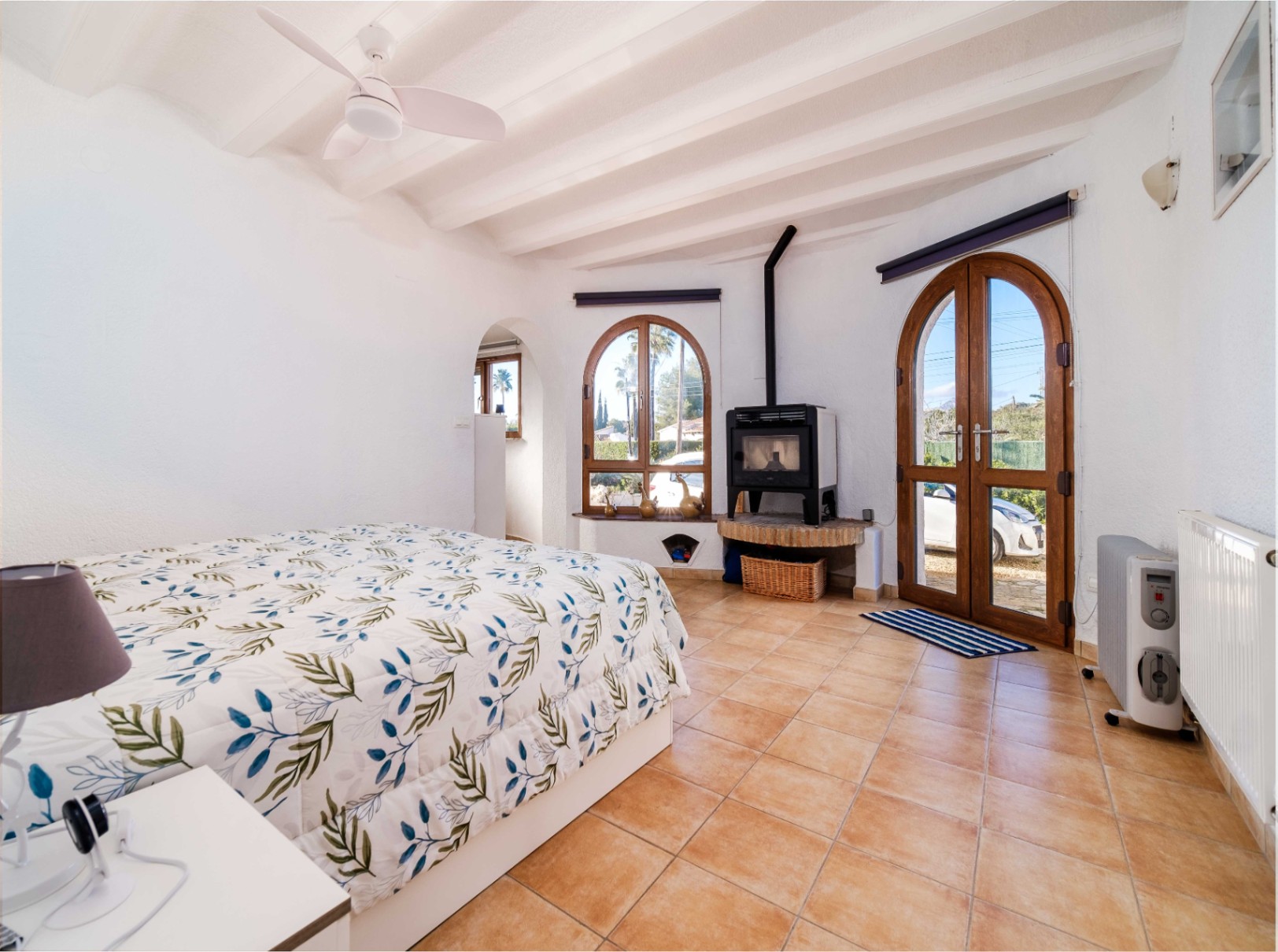 Helles Schlafzimmer mit weißer Balkendecke, Terrakottaboden und einem Holzofen. Rundbogentüren aus Holz führen direkt ins Freie und lassen viel Tageslicht herein.