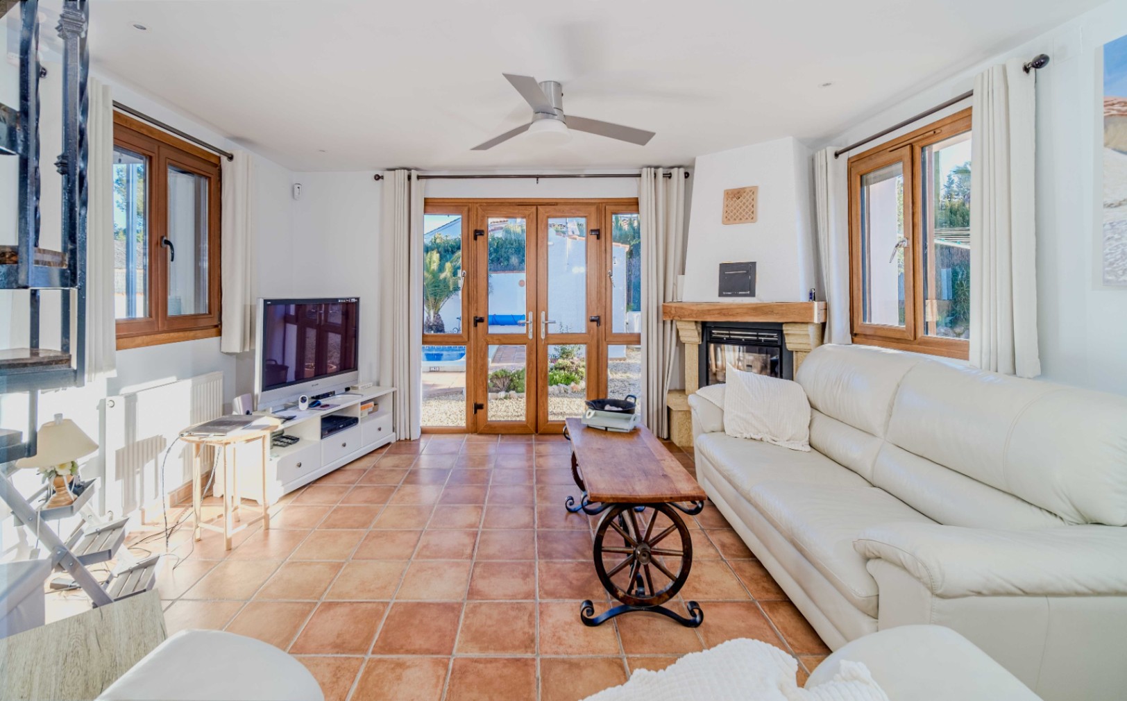 Helles Wohnzimmer mit Terrakottaboden, rustikalem Kamin und weißen Ledersofas. Große Holz-Glastüren führen direkt zum Außenbereich mit Pool.