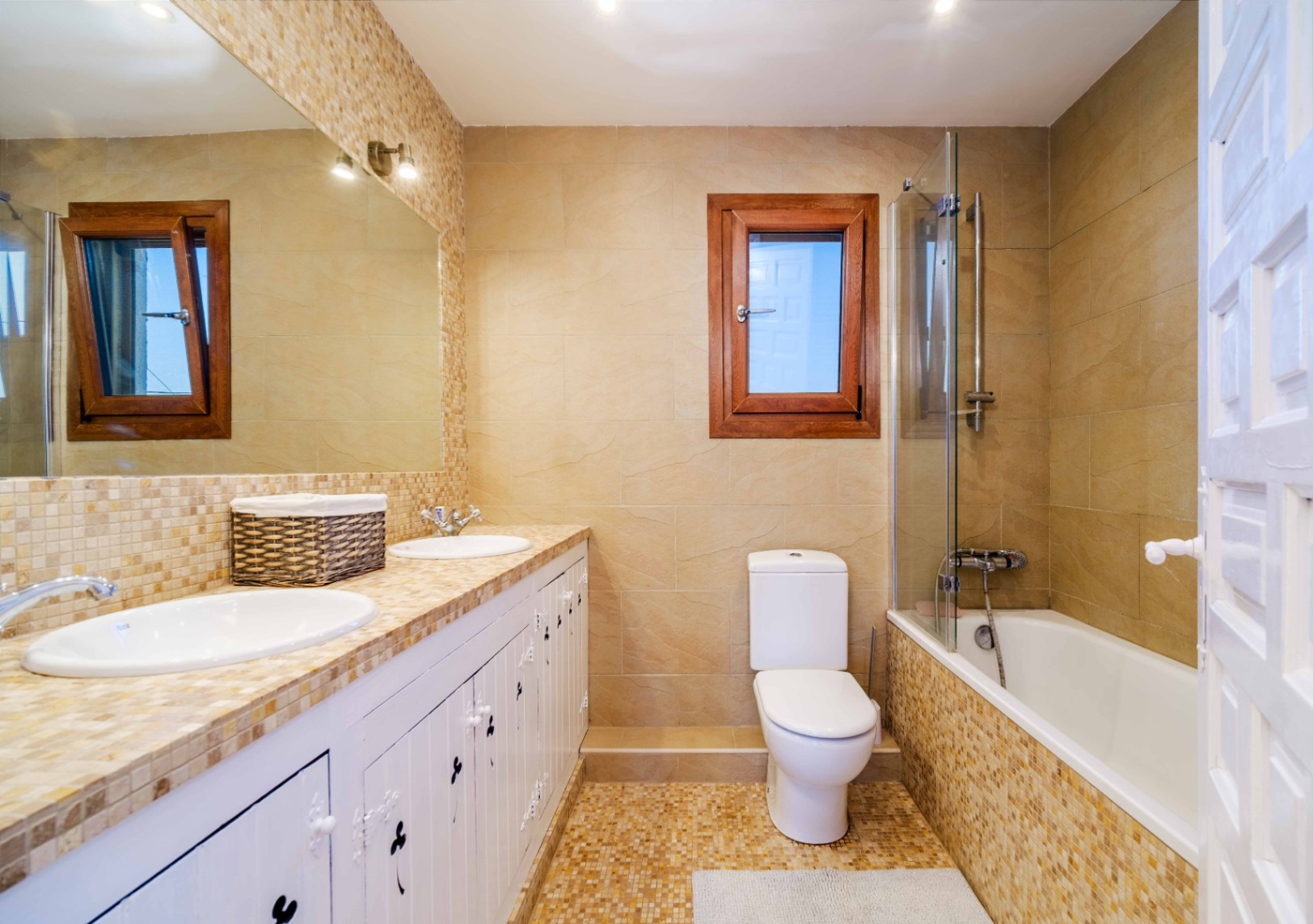 Komplettes Badezimmer mit Doppelwaschtisch auf Mosaik-Arbeitsplatte, weißen rustikalen Schränken und einer Badewanne mit Glastrennwand und erdfarbenen Fliesen.