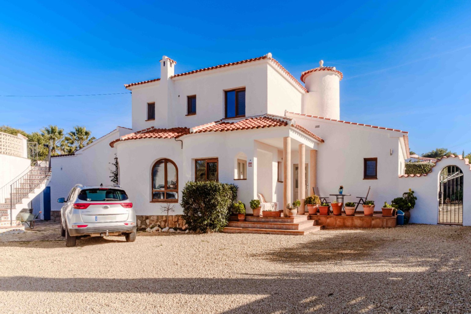 Mediterrane Villa mit weißer Putzfassade, Terrakotta-Ziegeldach, Säulenveranda und großzügiger Kiesauffahrt für mehrere Fahrzeuge.