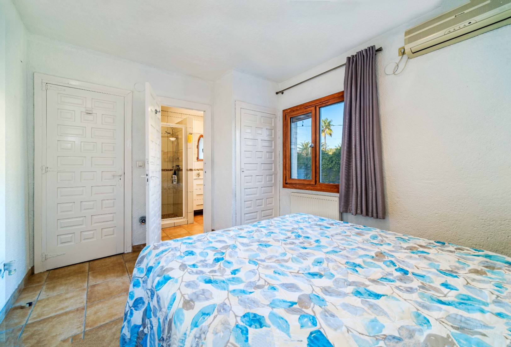 Schlafzimmer mit En-Suite-Bad, weißen Kassettentüren, Fliesenboden und Holzfenster. Inklusive wandmontierter Klimaanlage und Heizkörper unter dem Fenster.