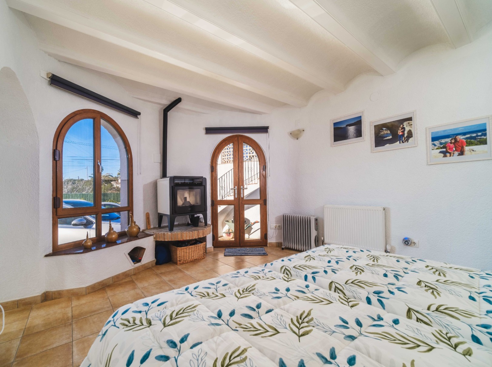 Schlafzimmer mit weißer Balkendecke, Kaminofen auf gemauertem Sockel und bogenförmigen Holzfenstern und -türen für viel Tageslicht.