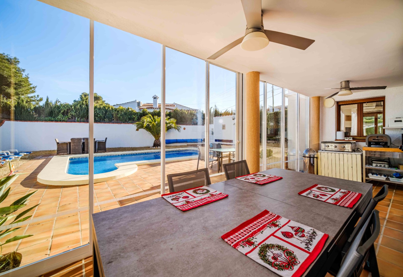 Verglaste Terrasse mit Essbereich, Deckenventilatoren und Außenküche, mit direktem Blick auf den privaten Salzwasserpool und den Garten.