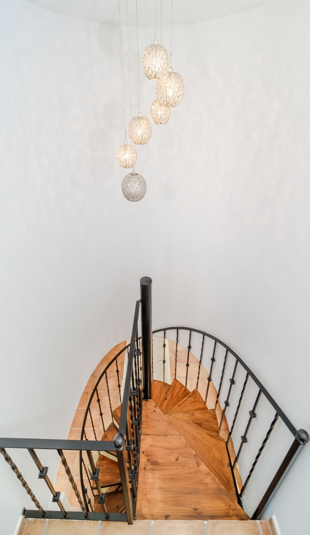 Wendeltreppe mit Holzstufen und schwarzem Schmiedeeisengeländer, beleuchtet durch eine Gruppe perforierter Kugelleuchten vor schlichten weißen Wänden.