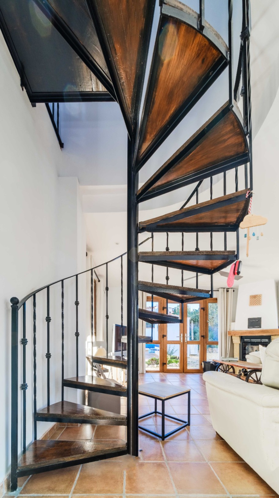 Wendeltreppe mit schwarzem Metallgestell und dunklen Holzstufen, integriert in einen Wohnraum mit Terrakottafliesen und direktem Gartenzugang.