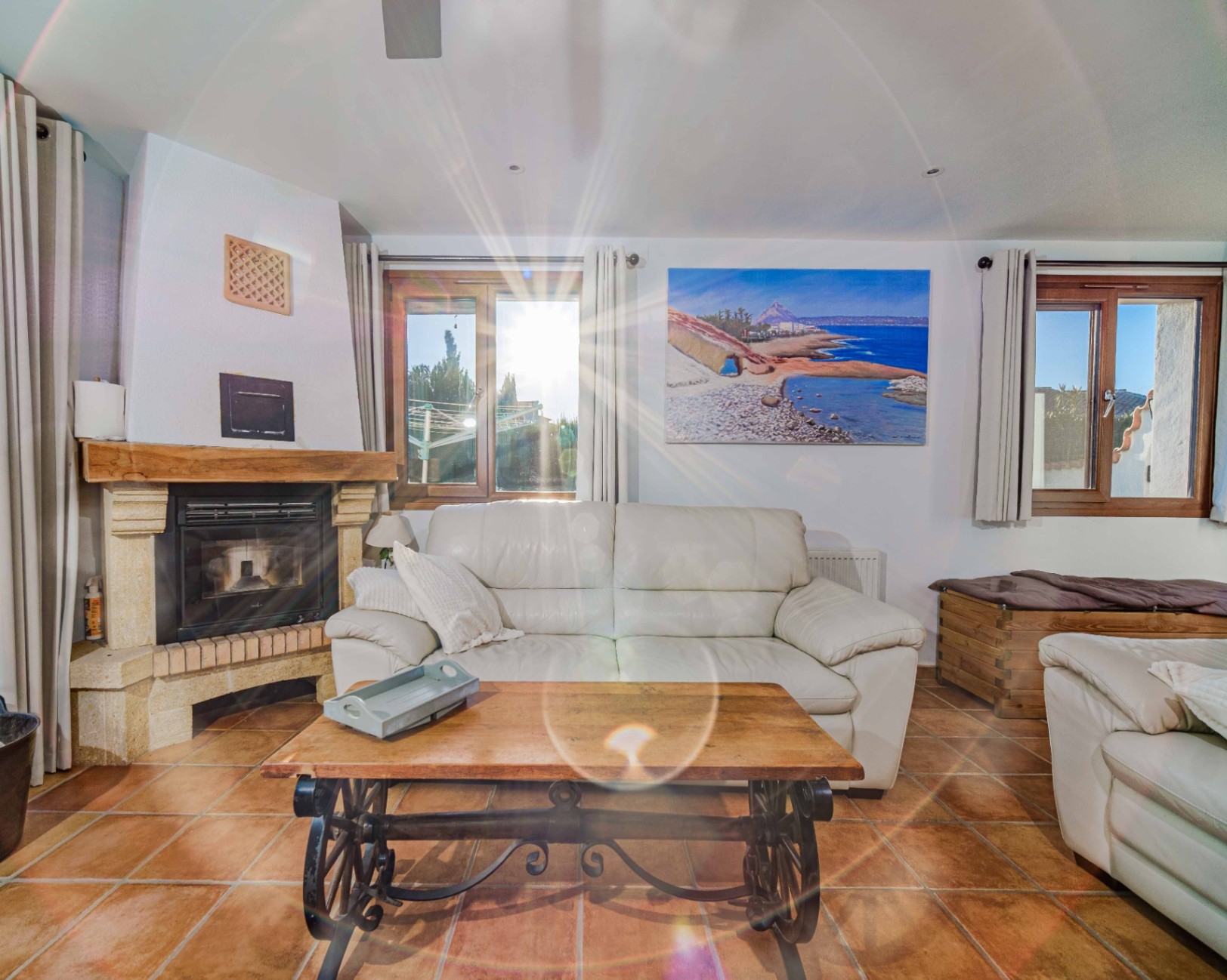 Wohnzimmer mit Natursteinkamin, Holzbalken, Terrakottaböden und weißen Ledersofas. Holzfenster sorgen für helles, natürliches Licht im gesamten Raum.