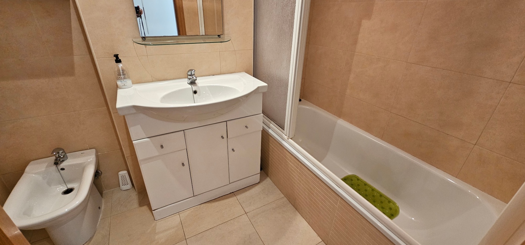 Badezimmer mit cremefarbenen Fliesen, ausgestattet mit einer Badewanne, einem Bidet und einem weißen Waschtisch mit Unterschrank.