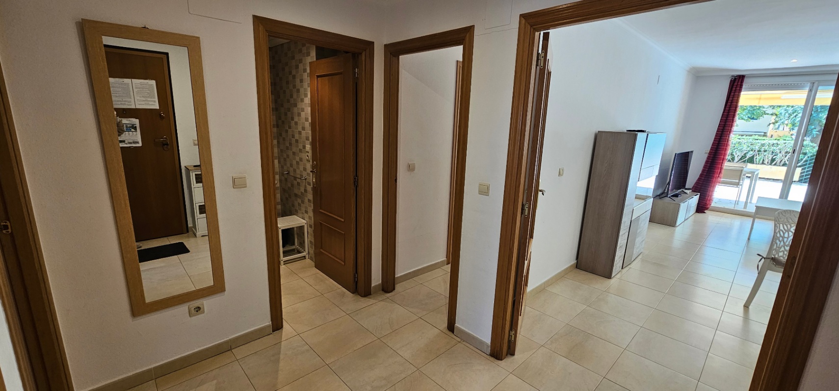 Flurbereich mit hellen Fliesenböden und Holztüren, der den Eingangsbereich mit einem Mosaikbad und dem zum Außenbereich offenen Wohnzimmer verbindet.